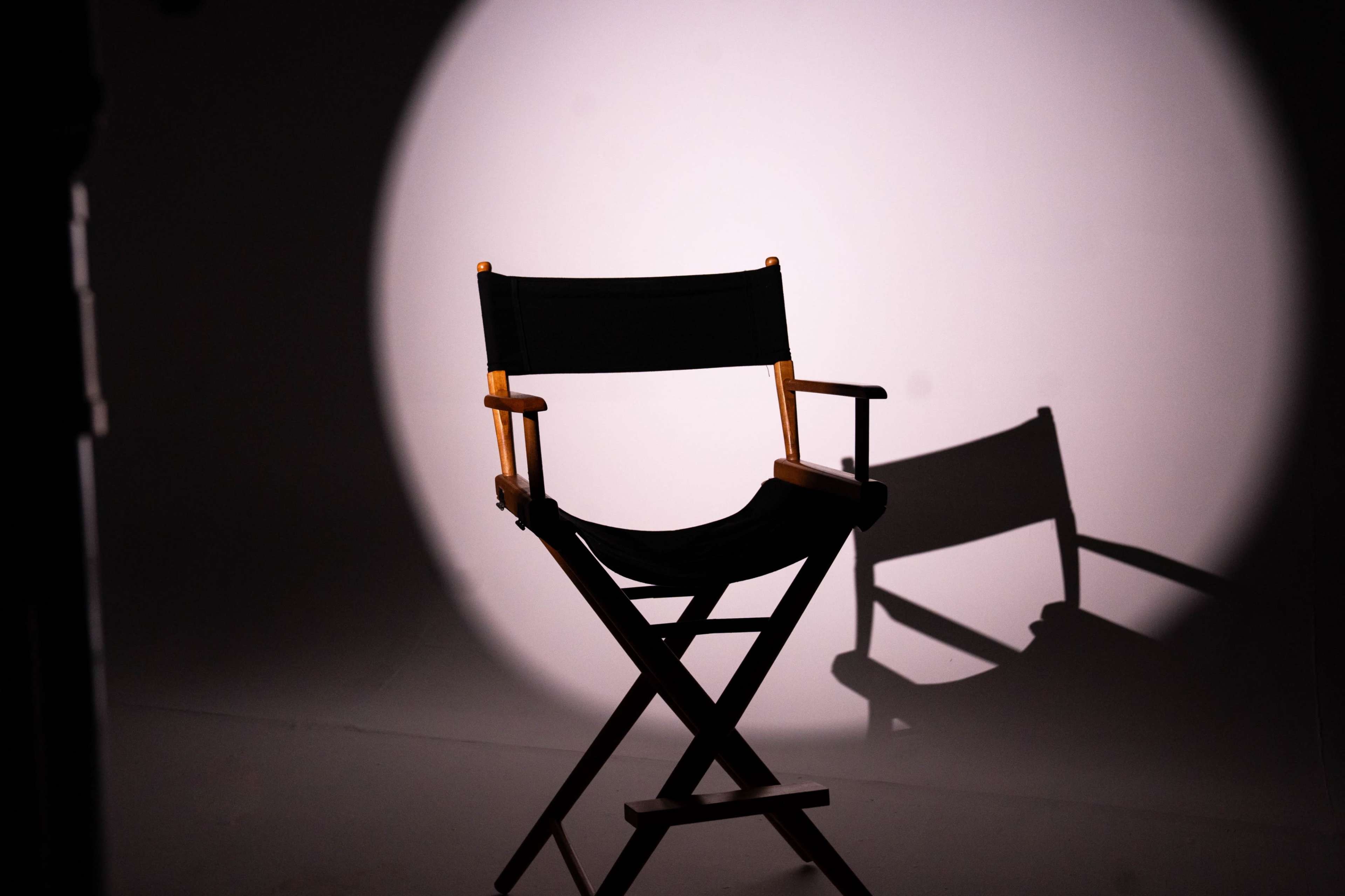 A director's chair casts a shadow in a spotlight against a plain backdrop.