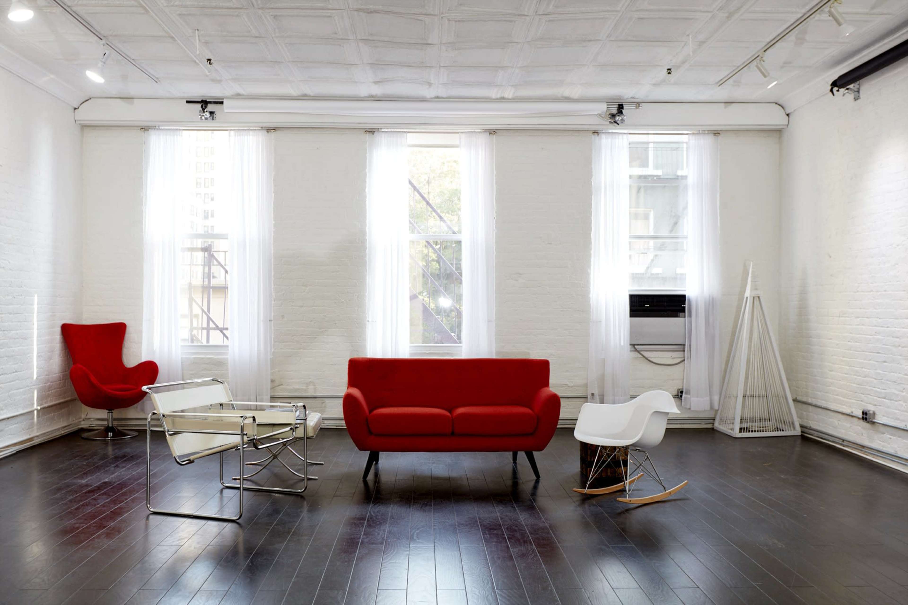 The image shows a modern, minimalist interior featuring a red sofa, a white rocking chair, a glass coffee table, and a white armchair, all arranged on a dark wooden floor with large windows letting in natural light.