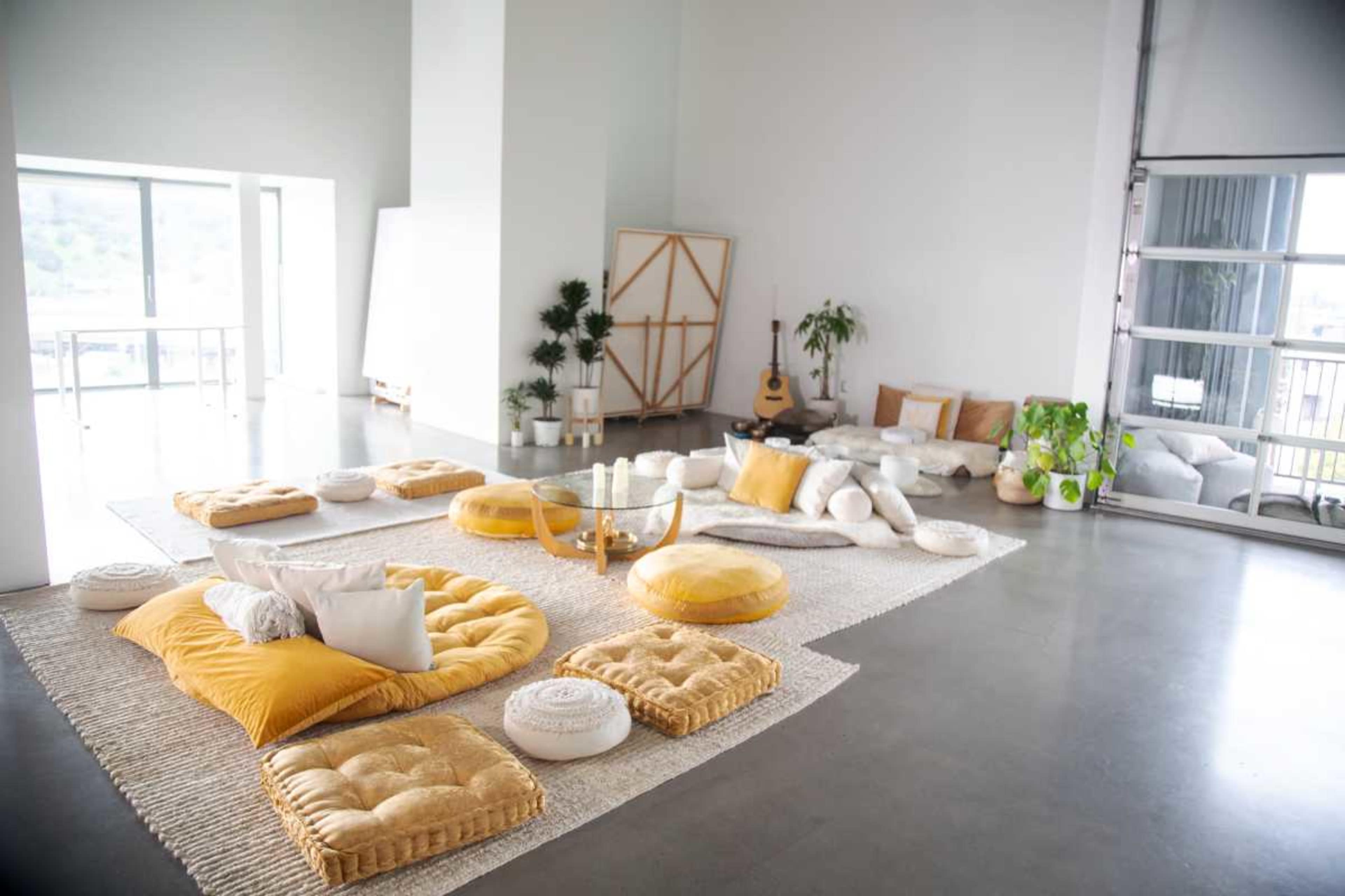 A spacious, minimalist living area furnished with yellow and white cushions arranged on a textured rug, alongside plants and a guitar.