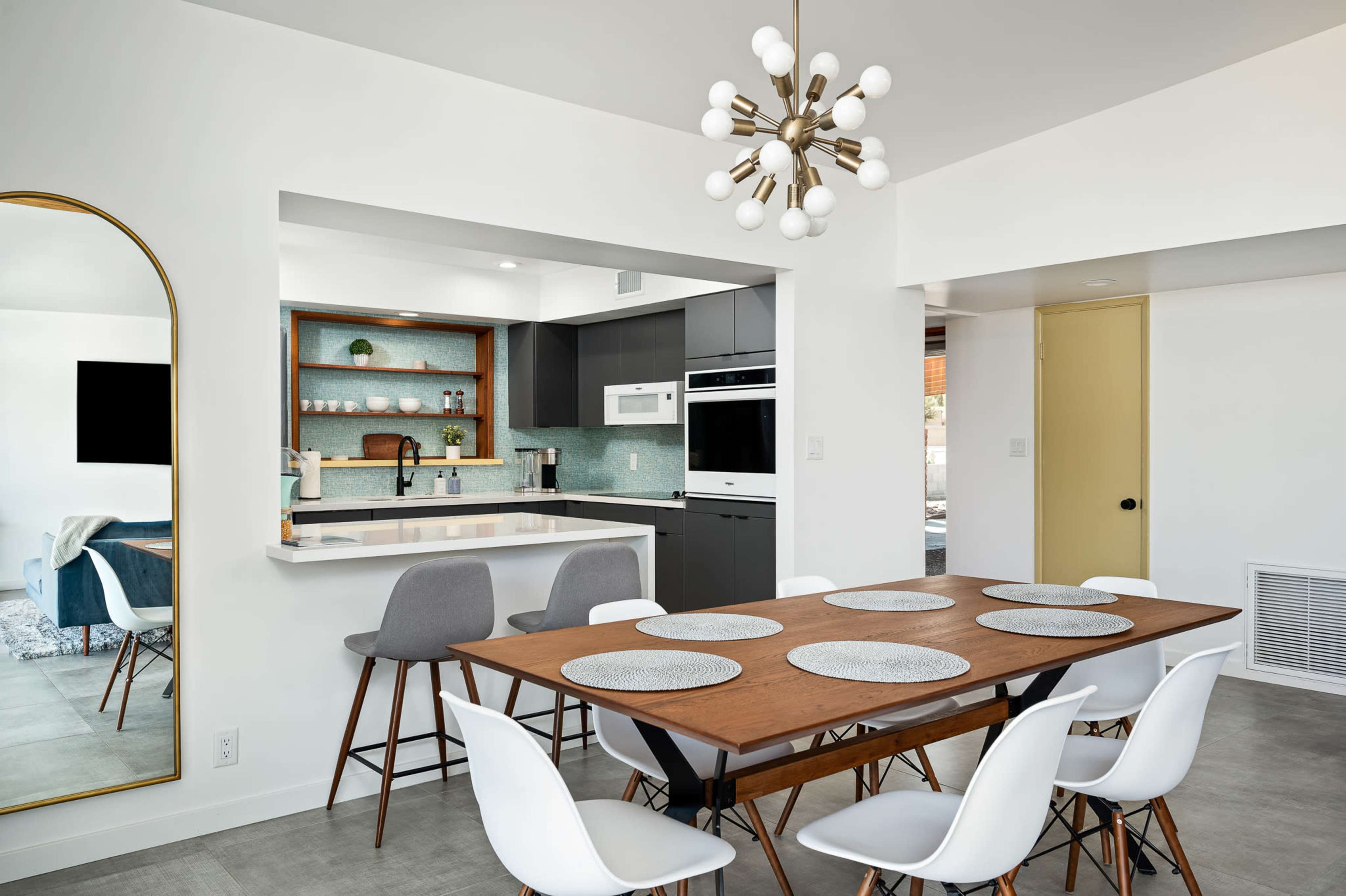 A modern dining area with a wooden table set for meals, surrounded by white chairs, with a kitchen featuring dark cabinetry and a light countertop in the background.
