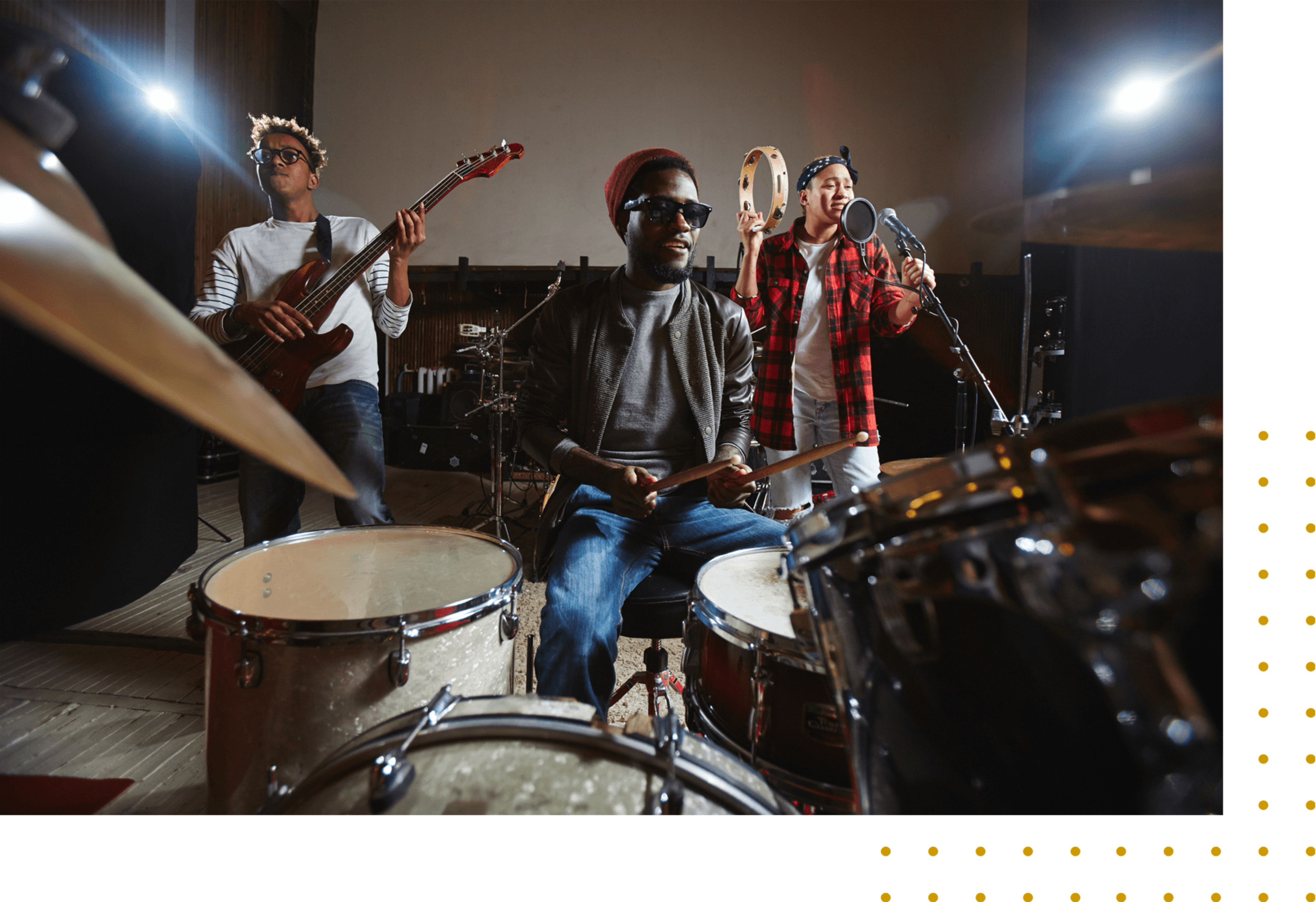 Three musicians perform together in a recording studio, with one playing the drums, another on bass, and a third singing into a microphone.