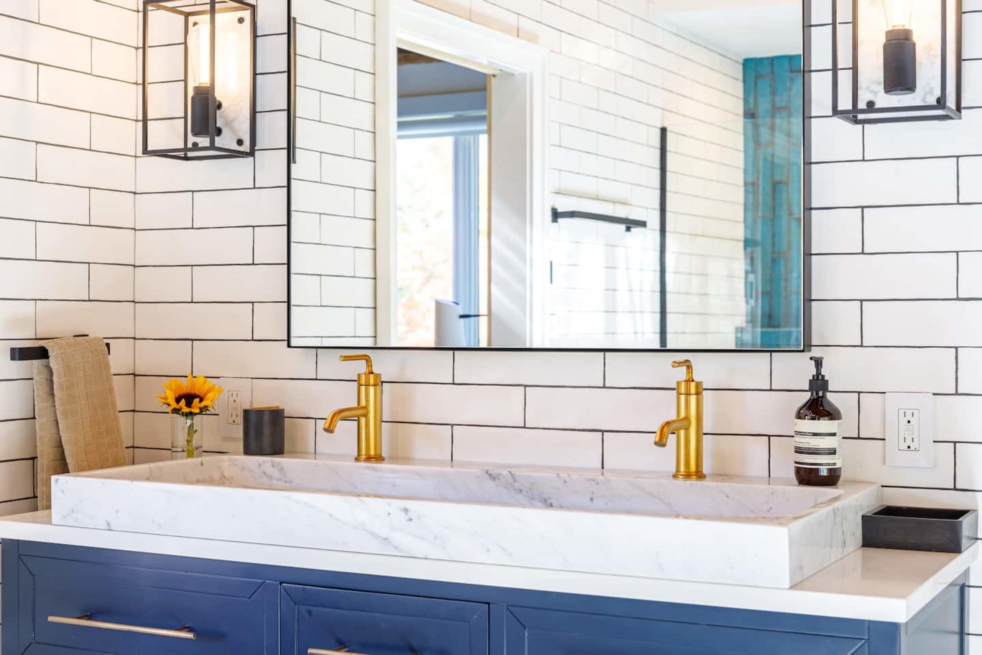 A bathroom sink area with a marble countertop, two gold faucets, a rectangular mirror, and tiled walls.