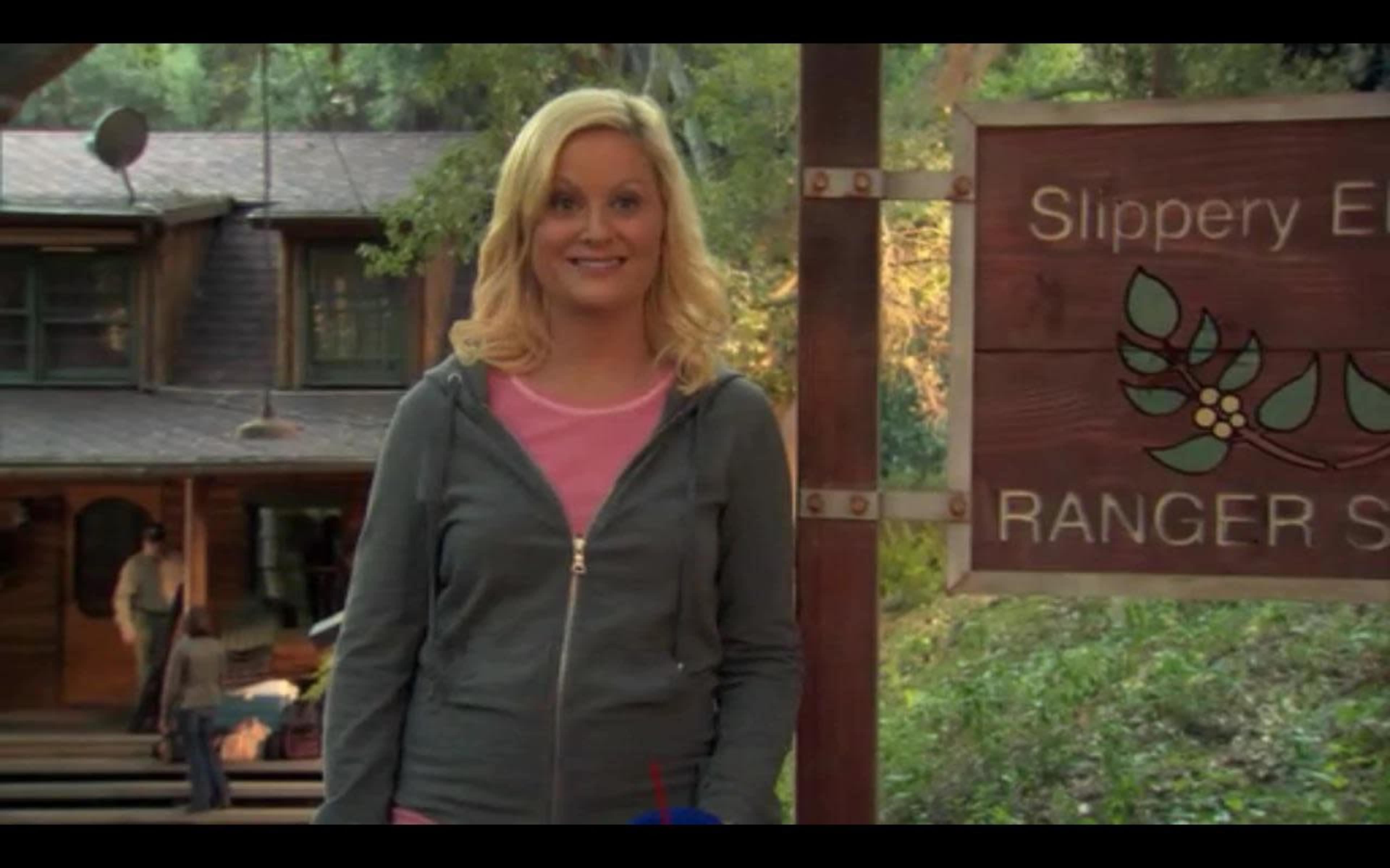 A woman stands outside a rustic ranger station with a smiling expression, holding a drink.