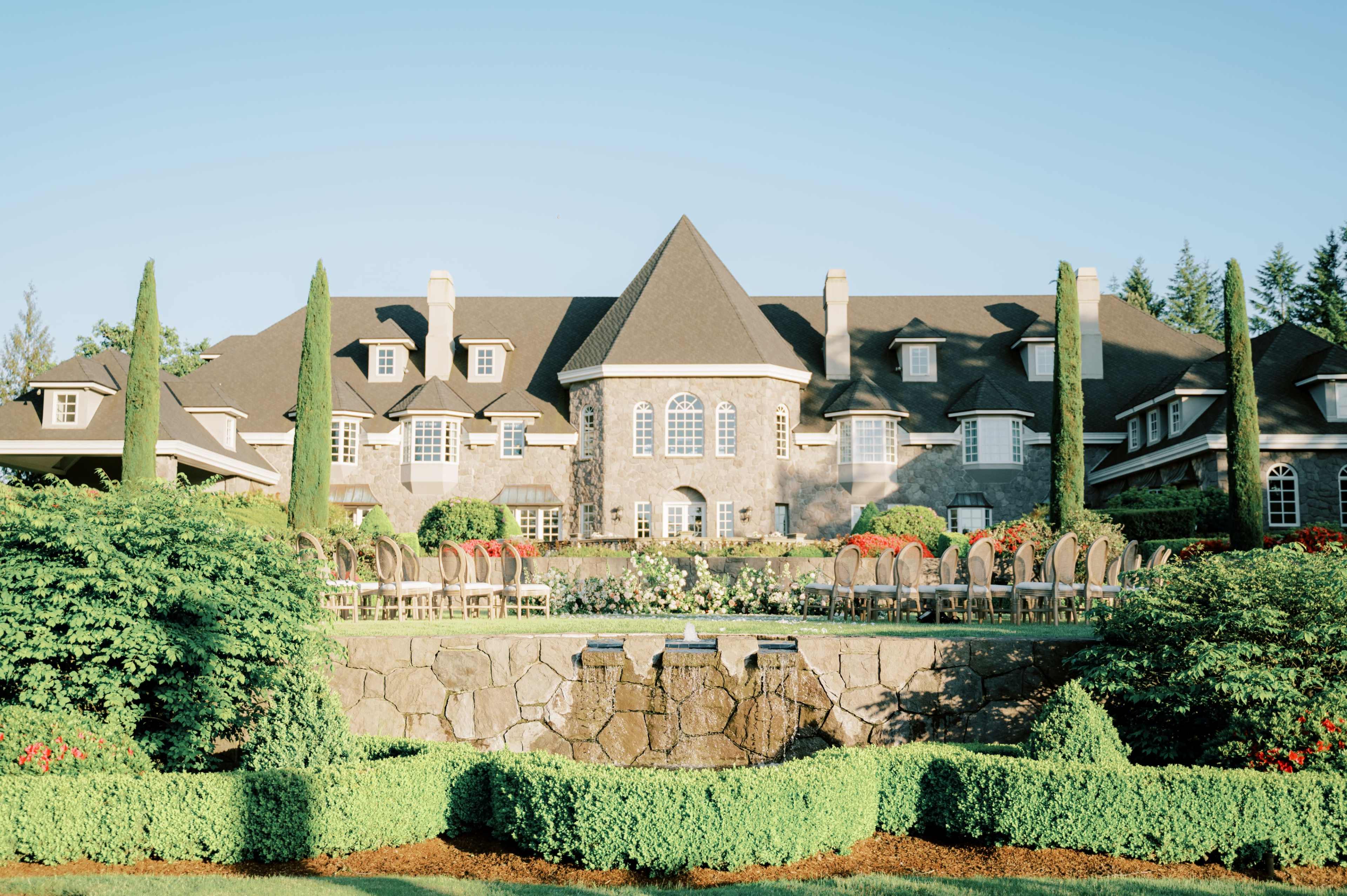 A large stone mansion with a steeply pitched roof, surrounded by well-maintained gardens and seating arranged on a terrace.
