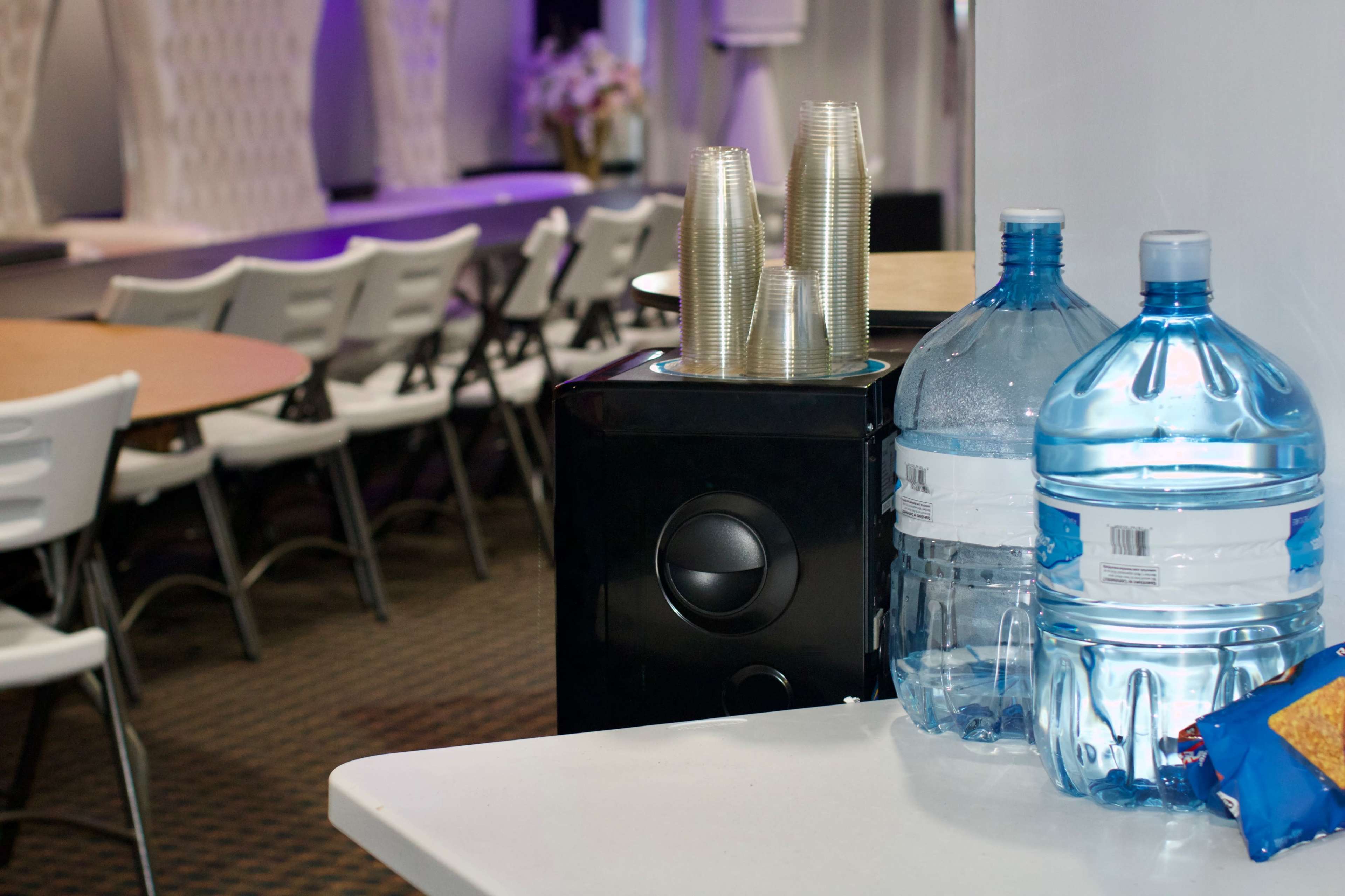 The image shows a table with two large water bottles, a stack of cups, and some snacks, with a backdrop of chairs and tables in a venue setting.