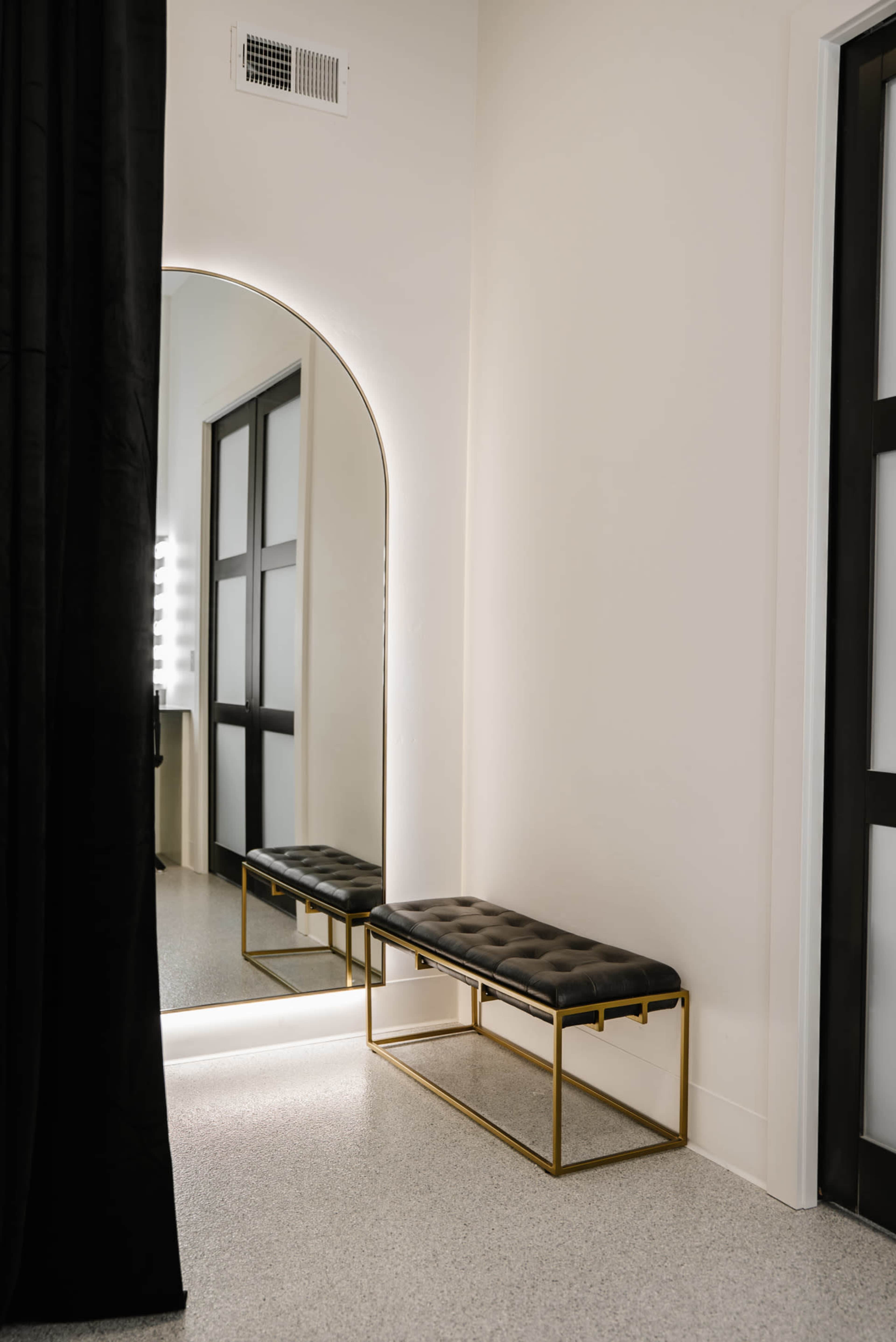 The image shows a cozy, minimalistic interior featuring a black tufted bench in front of a large mirror, with soft lighting and a curtain nearby.