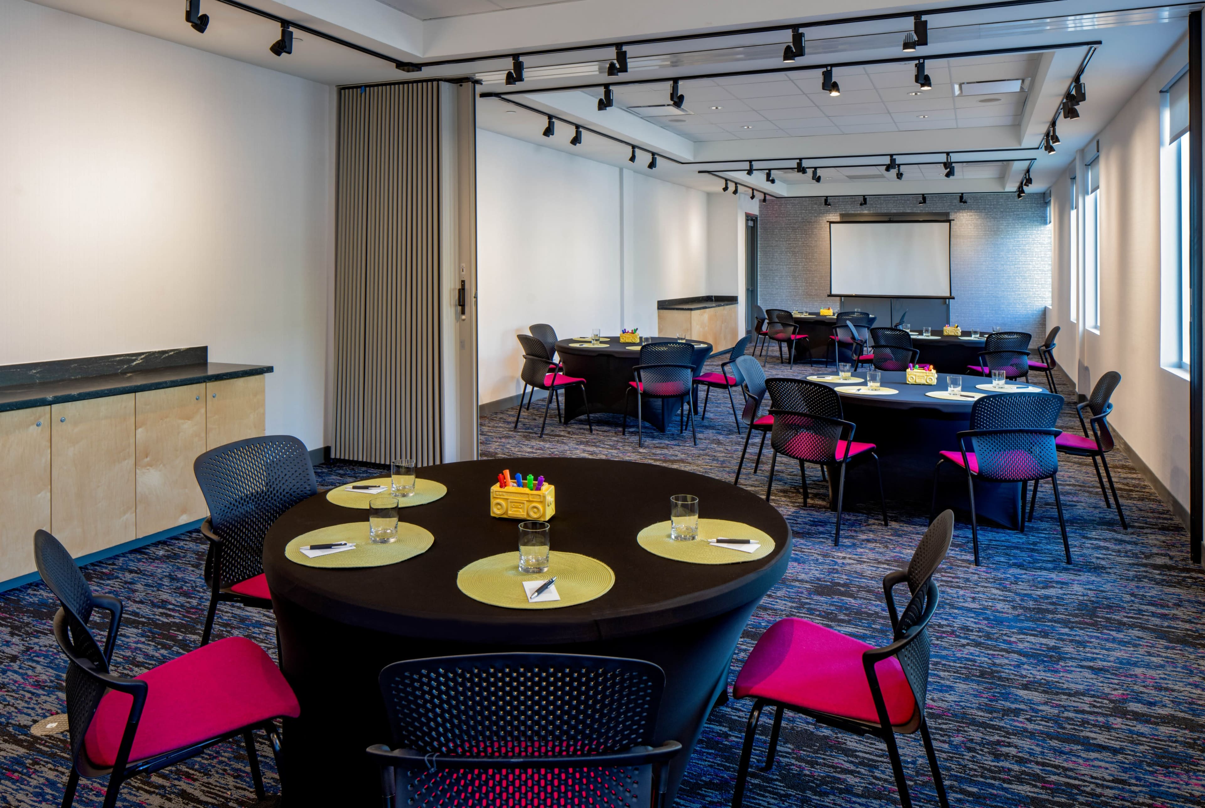 A conference room set up with round tables covered in black tablecloths and pink chairs, featuring a projector screen and a minimalist decor.