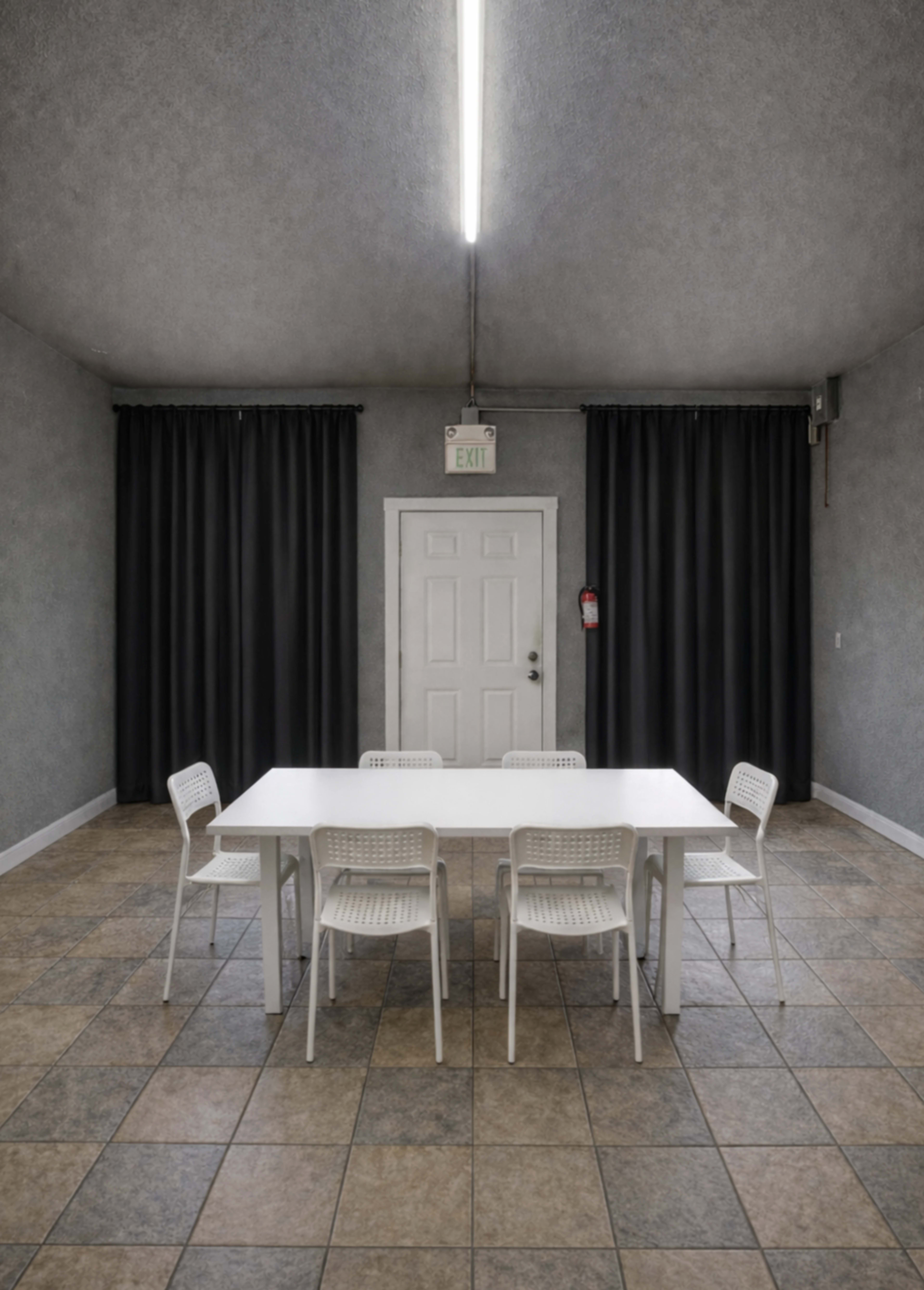 A rectangular white table is surrounded by six white chairs in a room with gray walls and black curtains.