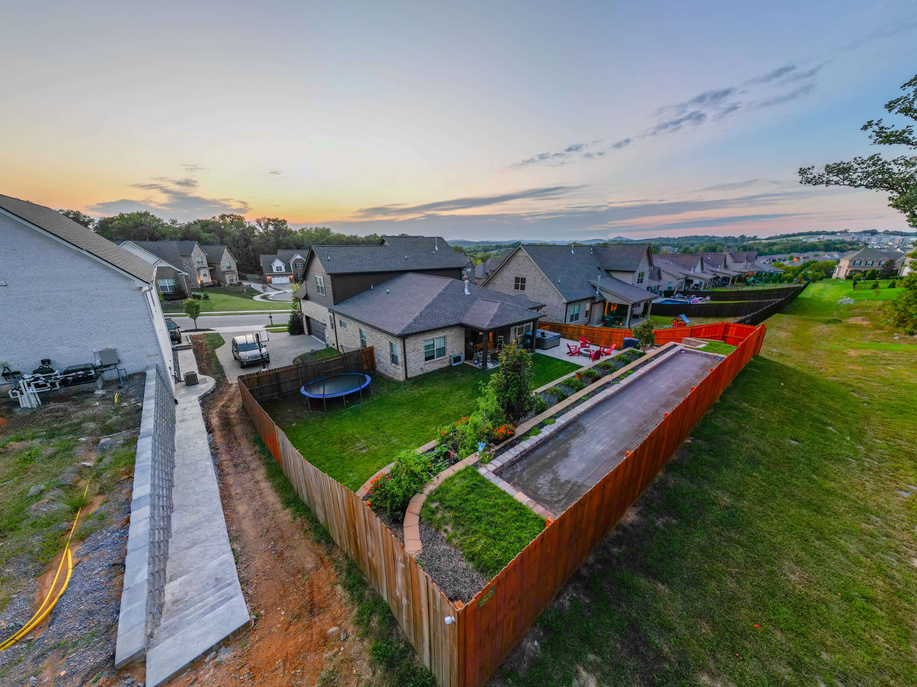 The image shows a suburban neighborhood with several houses, a backyard garden, a trampoline, and a view of a sunset in the background.