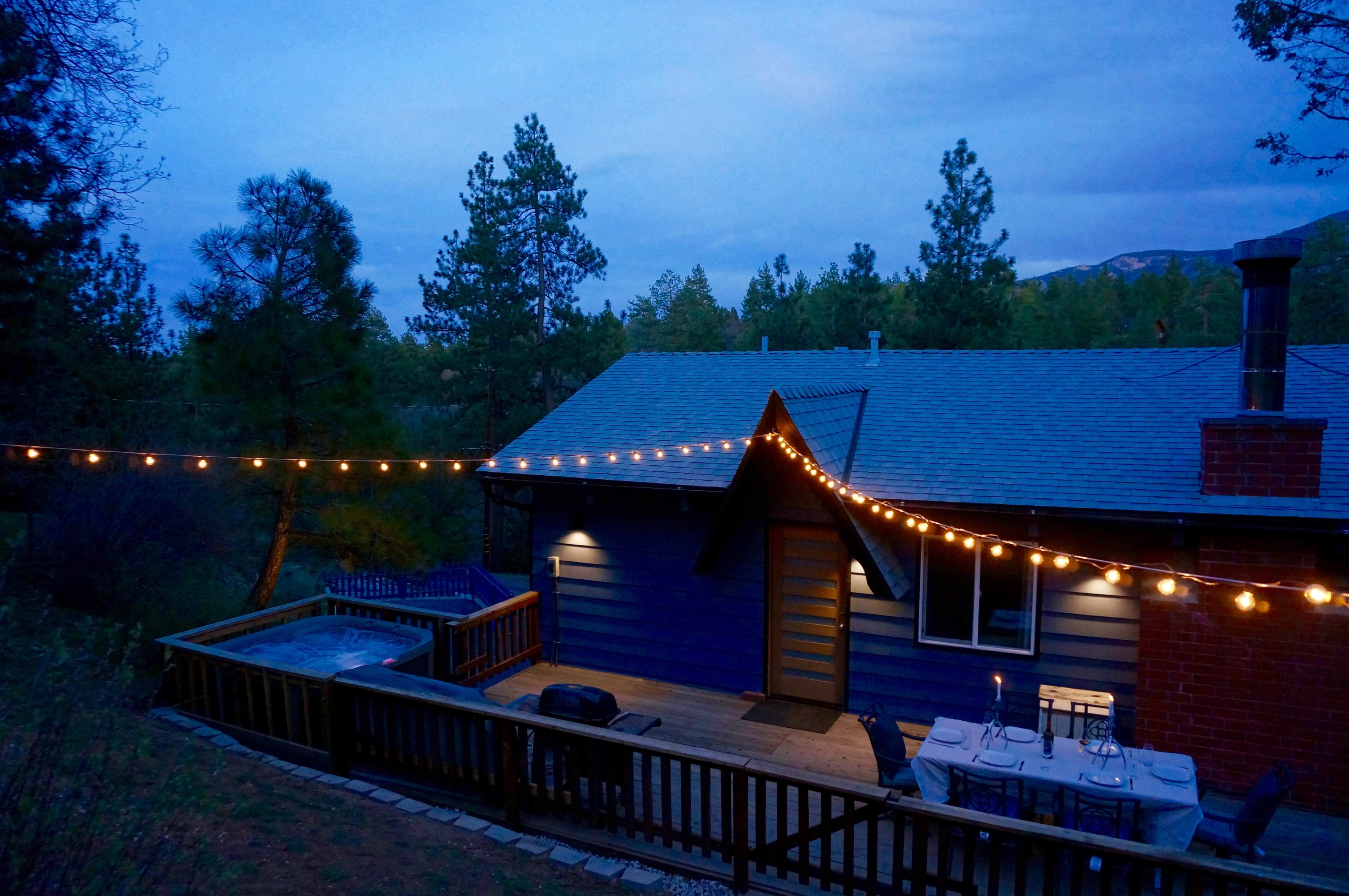 A cabin with string lights is situated near a hot tub on a wooden deck, surrounded by pine trees at dusk.