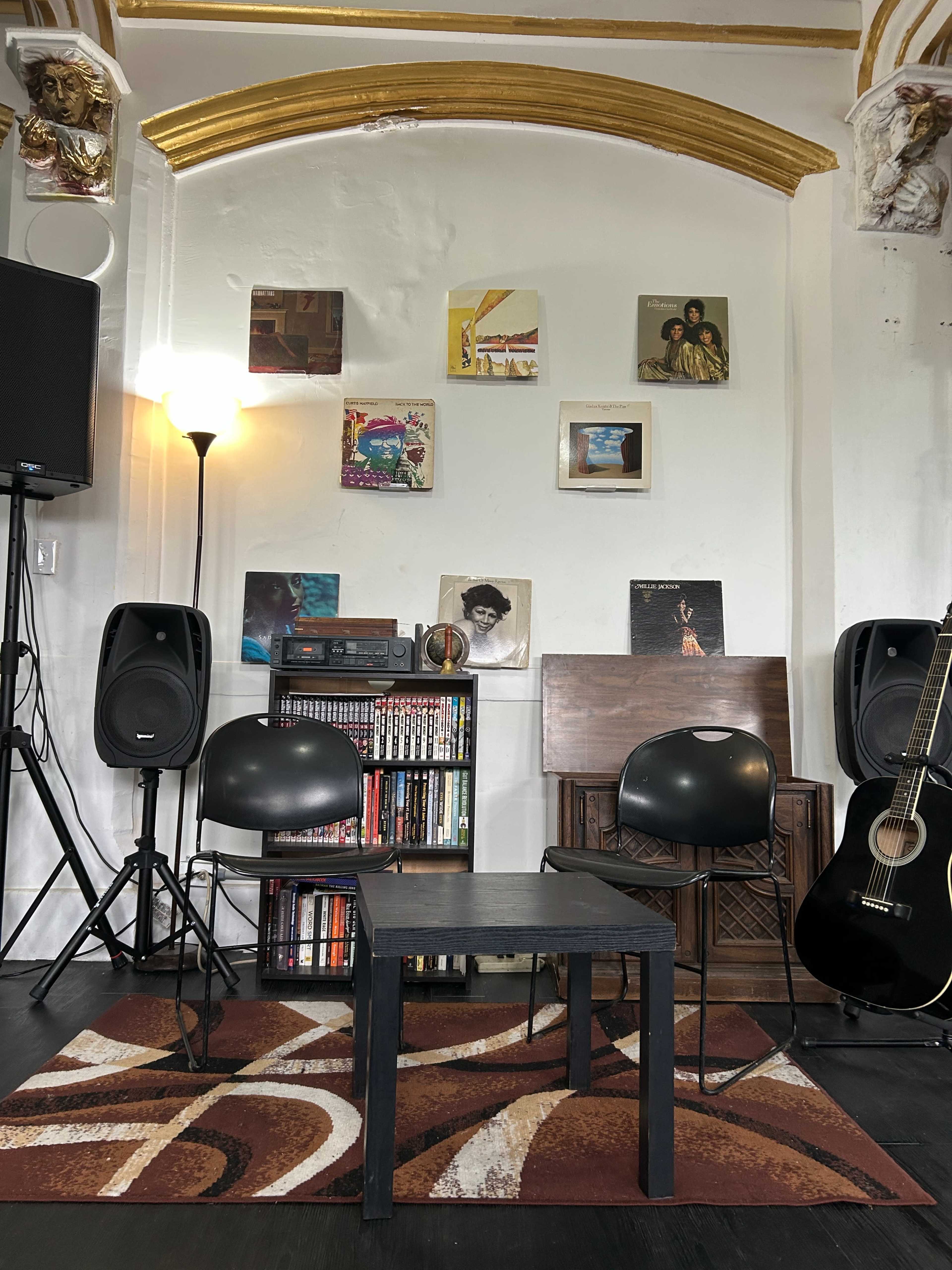 The image features a small stage setup with two black chairs, a table, speakers, an acoustic guitar, and a wall adorned with colorful album covers and artwork.