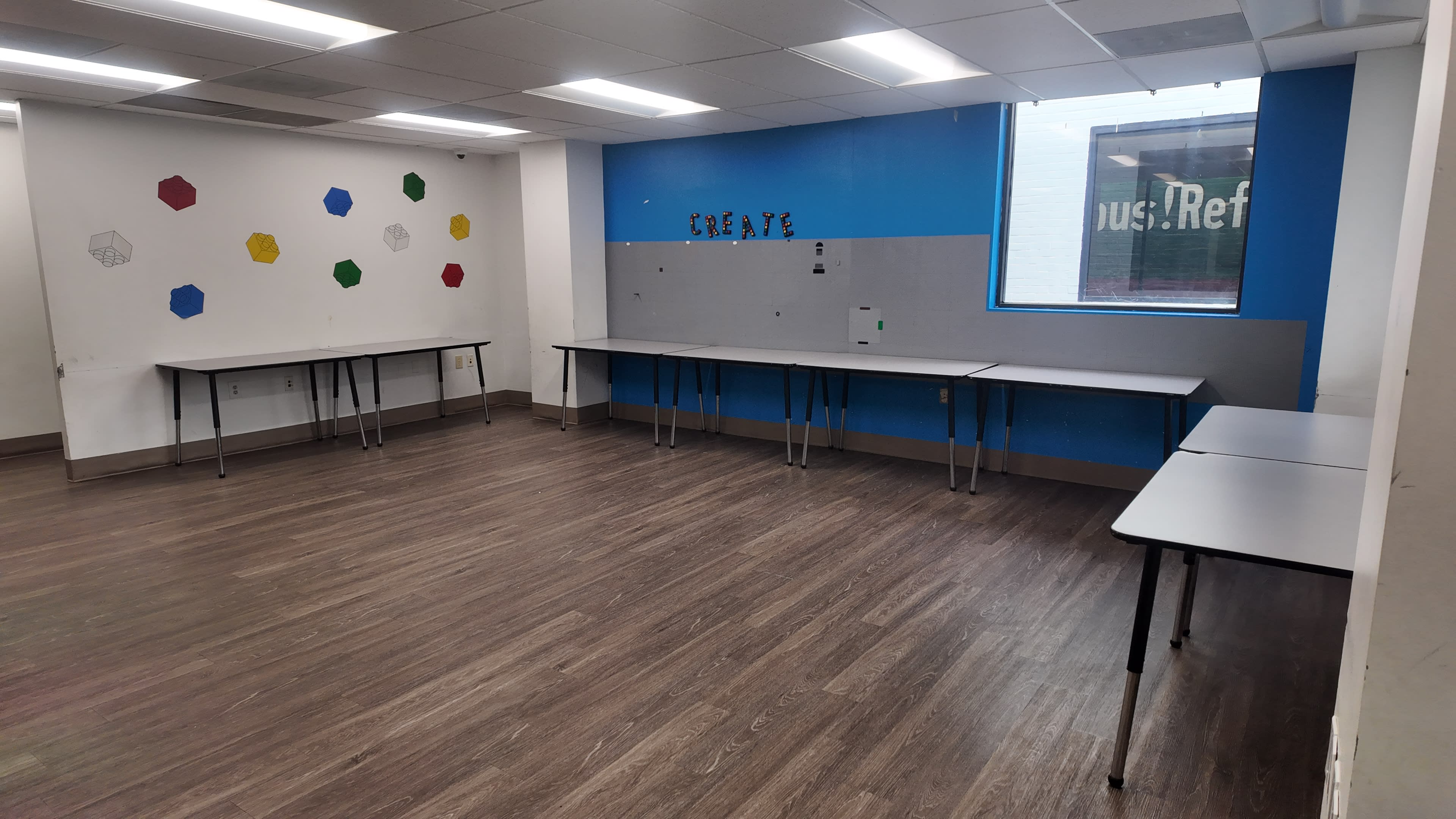 The image shows a spacious classroom with several tables arranged in a rectangular layout against walls painted in white and blue, adorned with colorful geometric shapes.