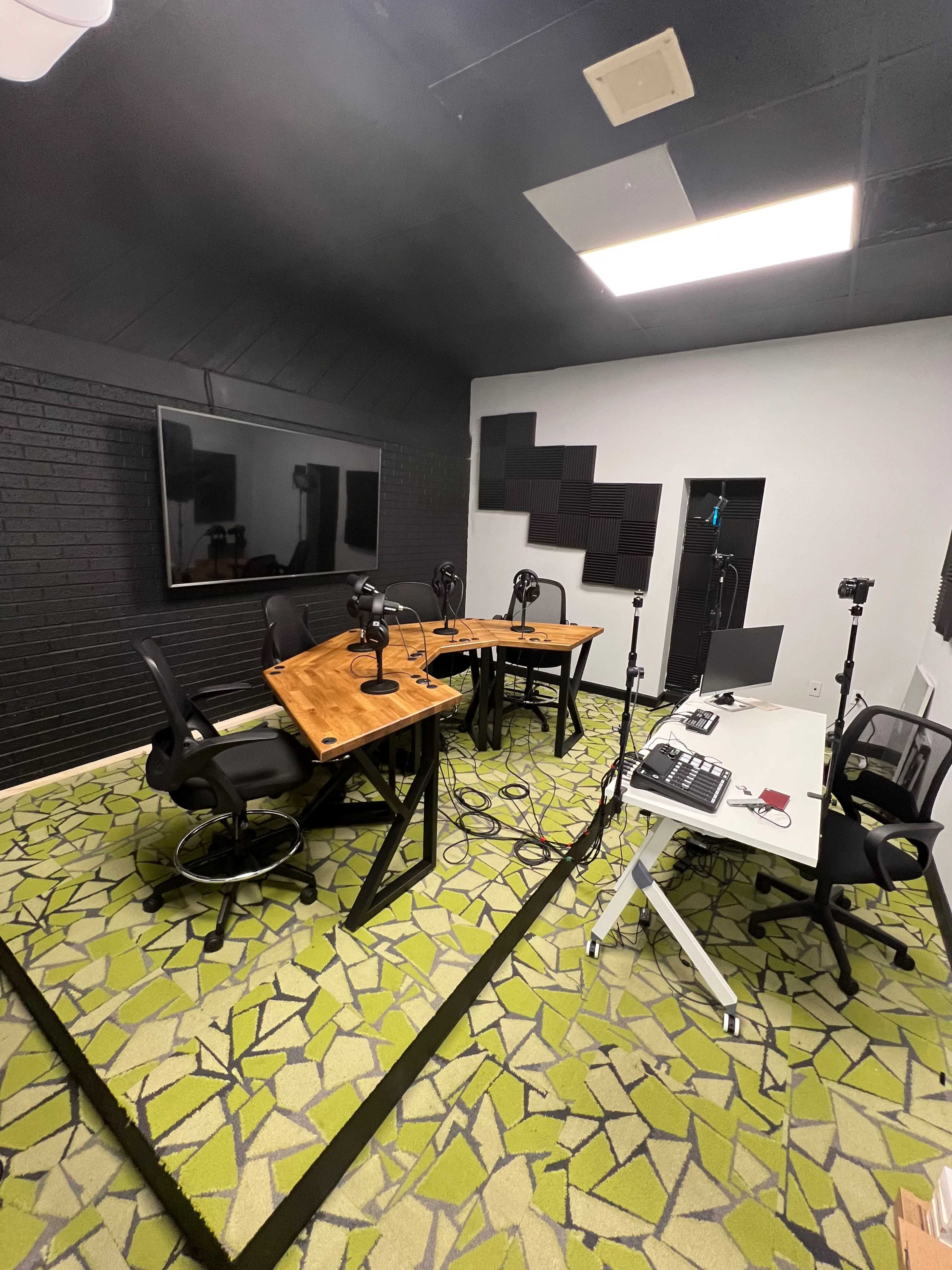 A recording studio with two wooden tables, each equipped with microphones, surrounded by sleek office chairs, and featuring a large screen on the wall and a desk with a computer.