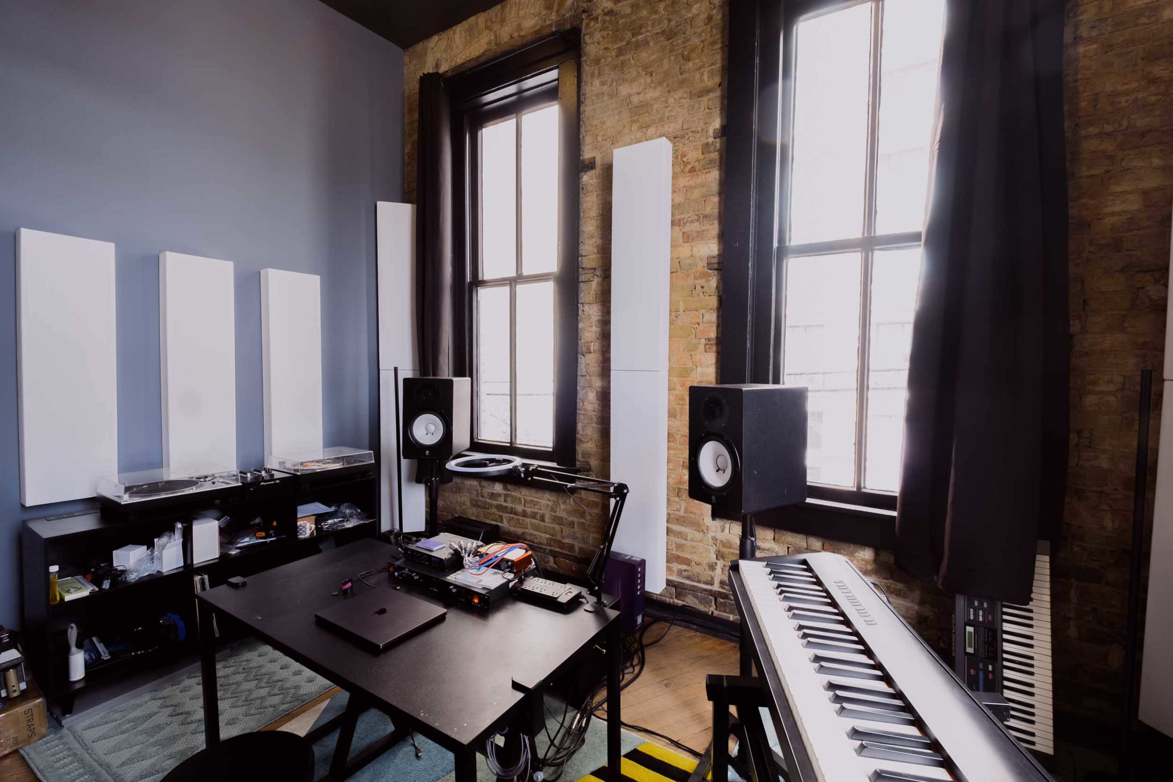 The image shows a music production studio featuring a black workstation, studio speakers, a keyboard, and large windows with natural light illuminating the brick walls.