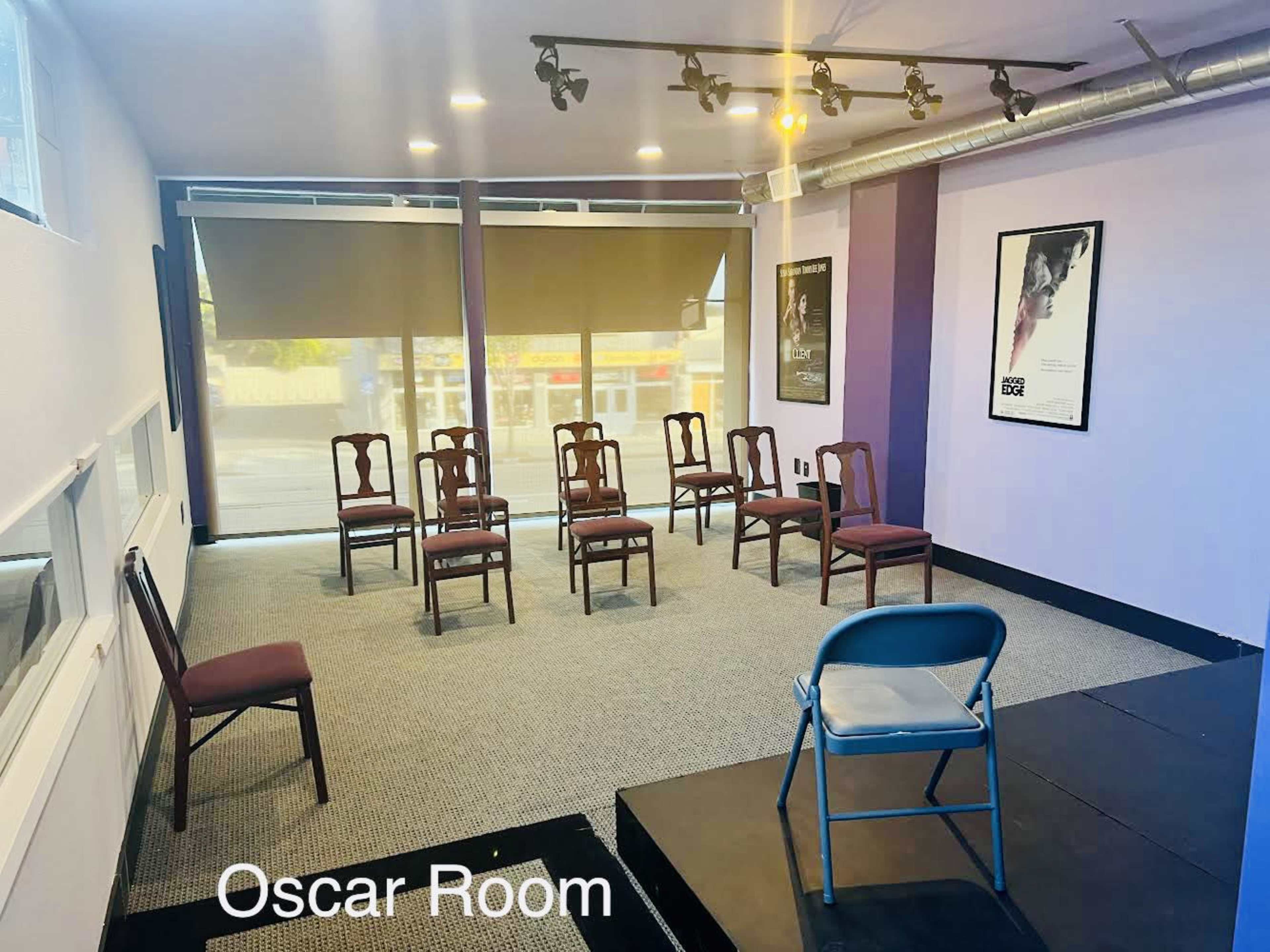 A meeting room with several wooden chairs arranged in a circle on a carpeted floor, featuring large windows and posters on the walls.