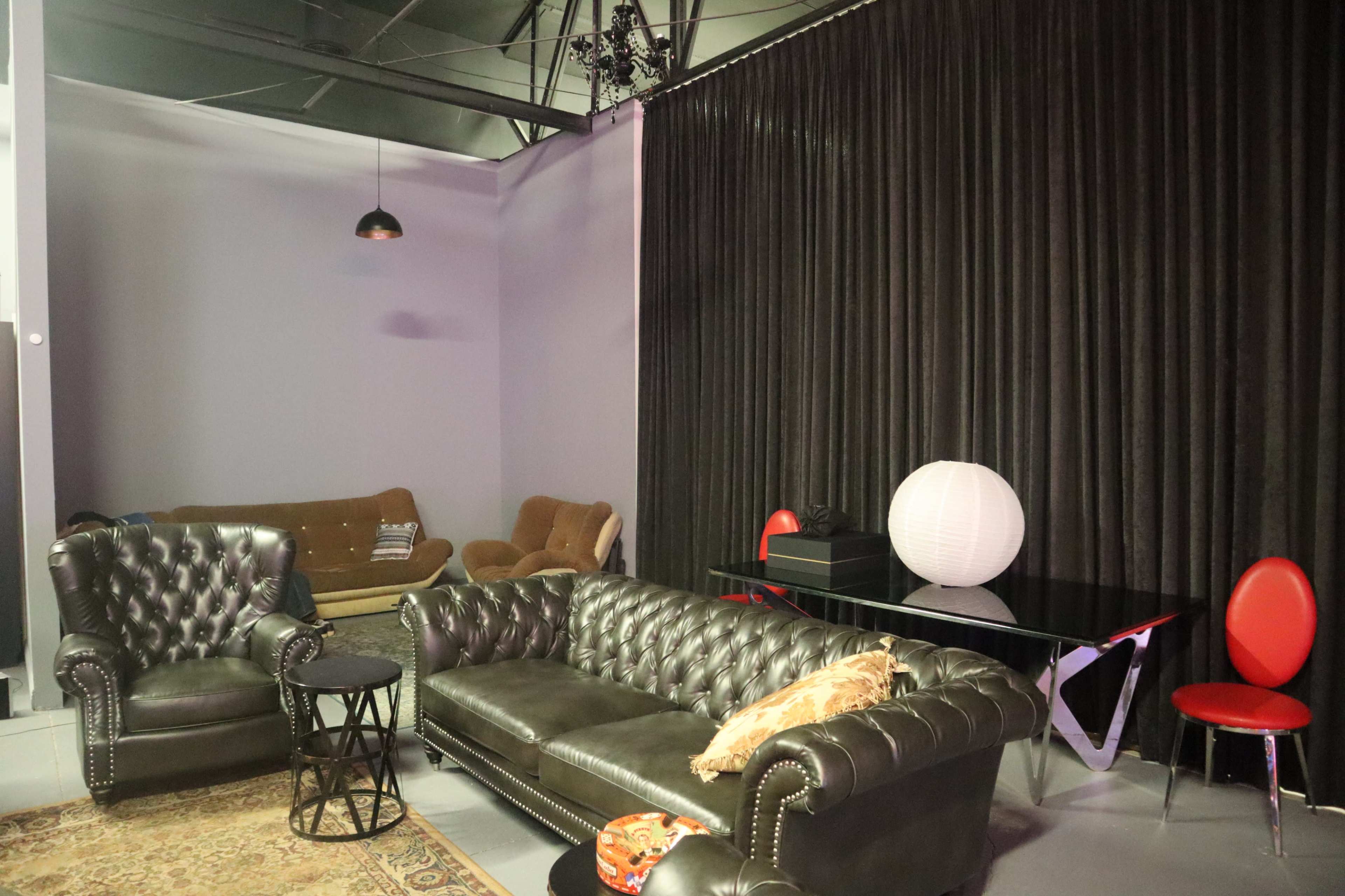 A modern lounge area with a black leather couch, two upholstered chairs, a round side table, and a white paper lantern on a black table, all set against a backdrop of black curtains.
