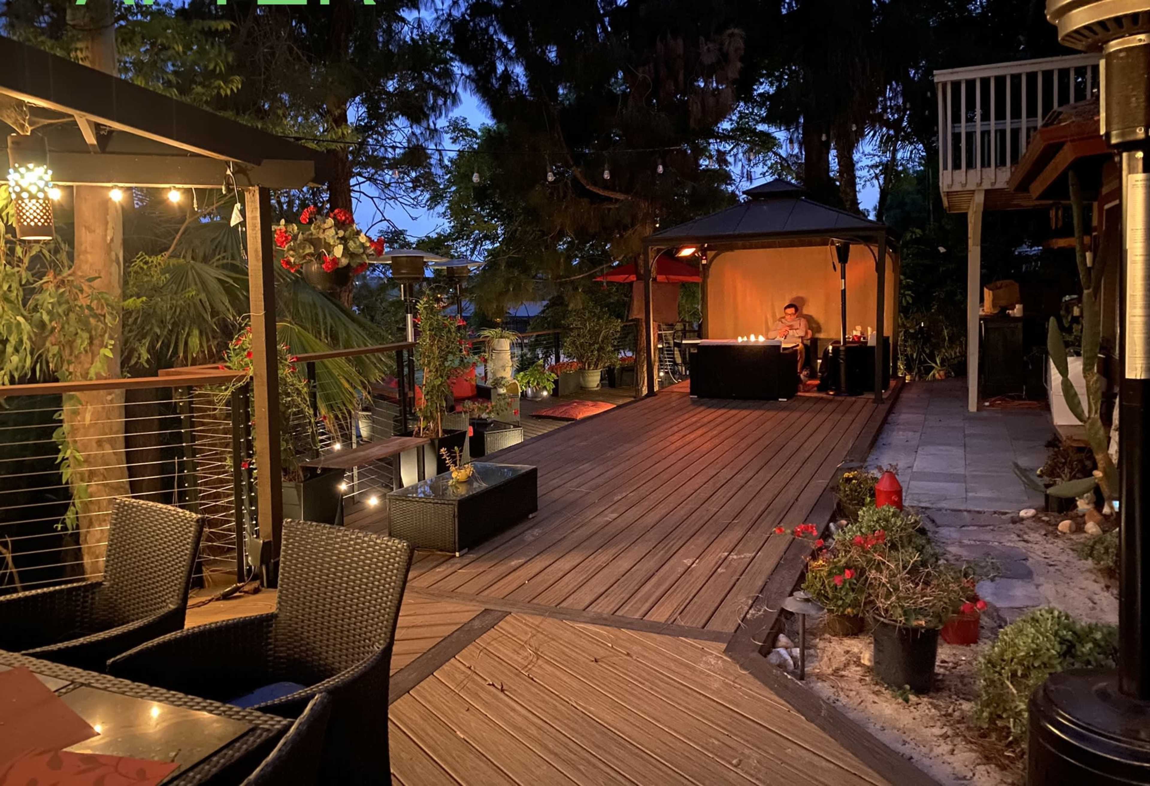 A wooden deck at dusk features a gazebo with a table set for dinner and potted plants along the walkway.