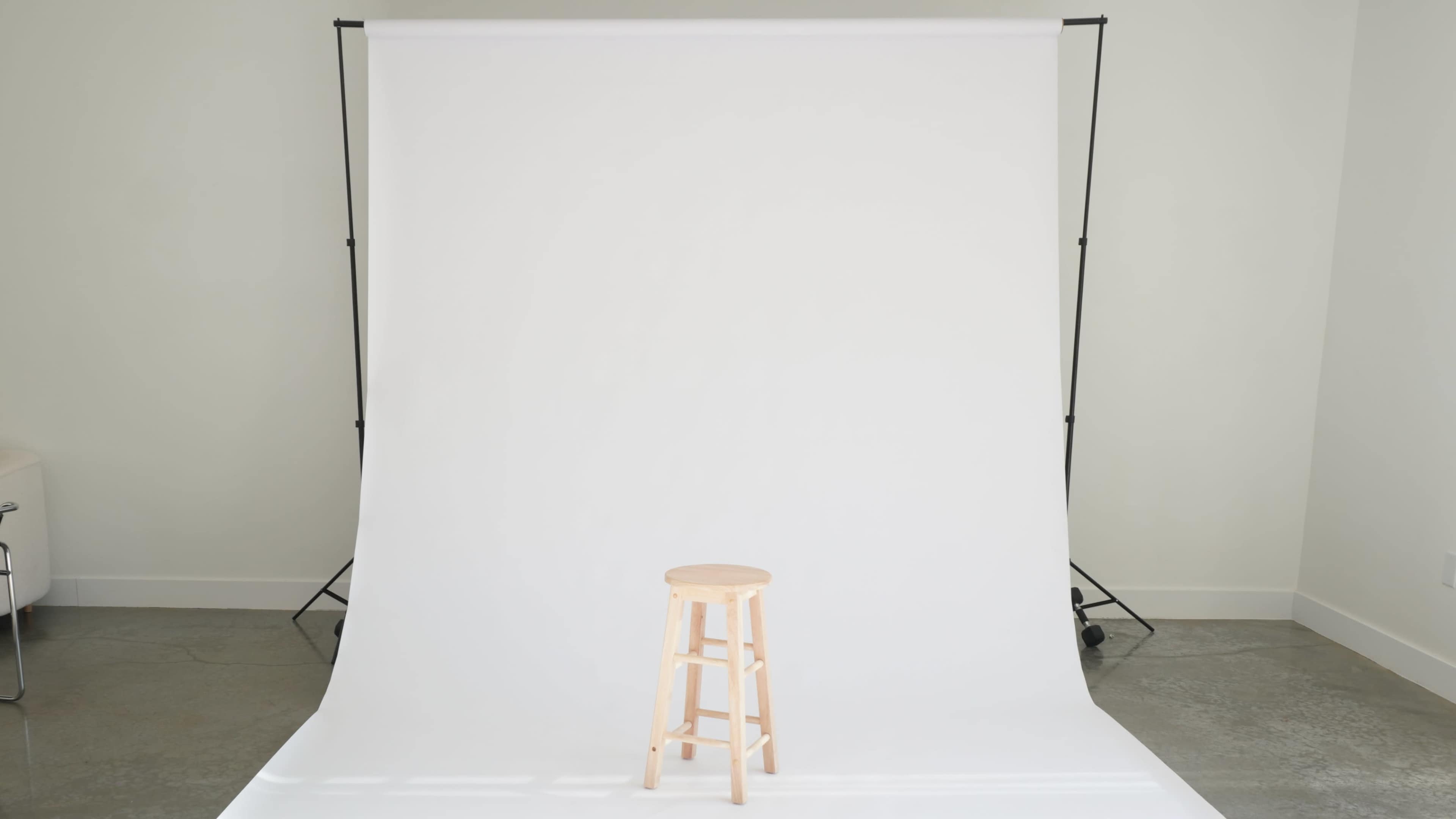 A wooden stool is positioned in front of a large blank white backdrop in a studio setting.