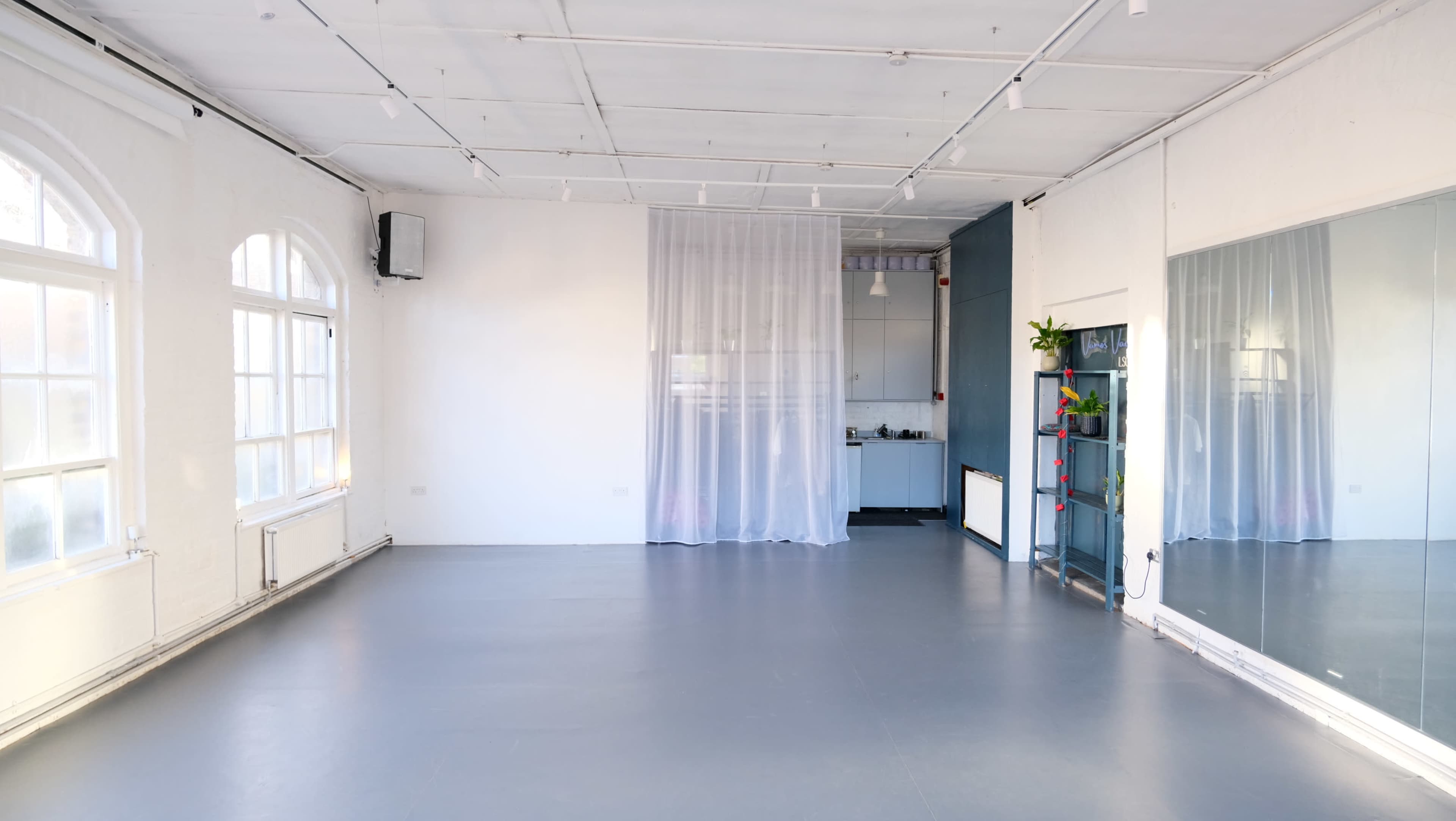 The image shows a spacious, empty studio with large windows, a partially separated kitchen area, and a mirror on one wall.