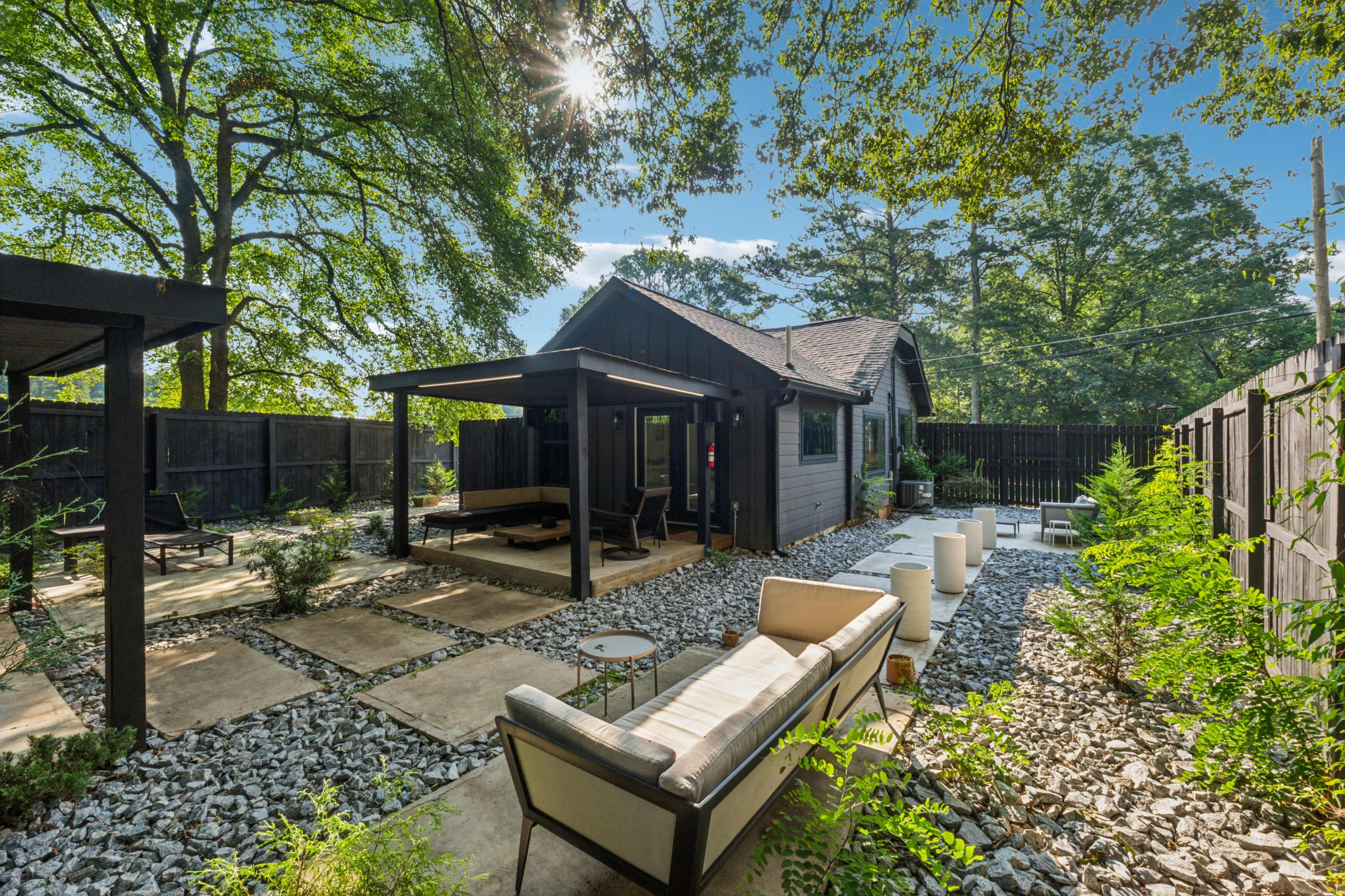 The image shows a landscaped outdoor area featuring a modern wooden house, gravel paths, and a seating arrangement under a shaded pergola.