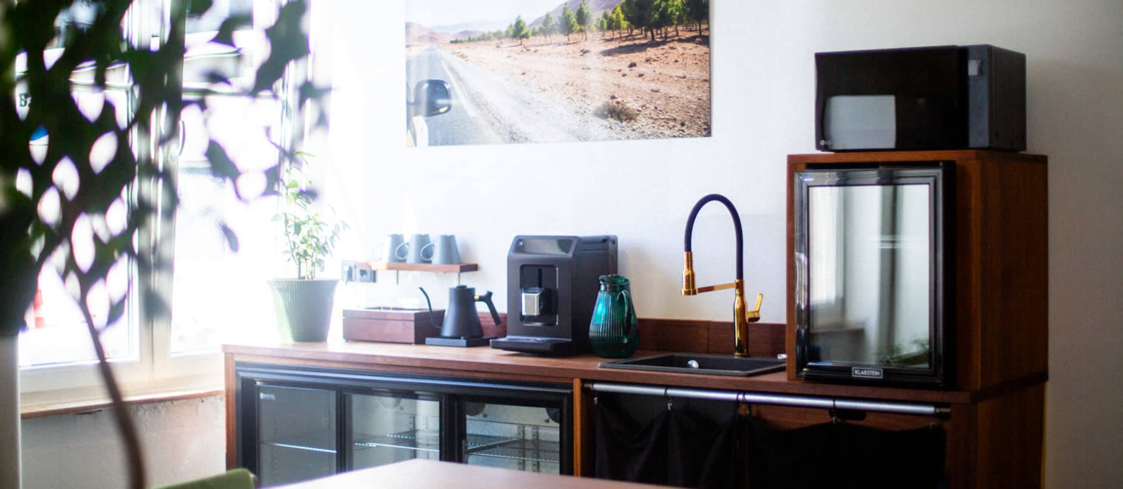 A kitchen corner features a refrigerator, a microwave, a coffee machine, and a plant on a wooden countertop next to a window with a landscape photo above.
