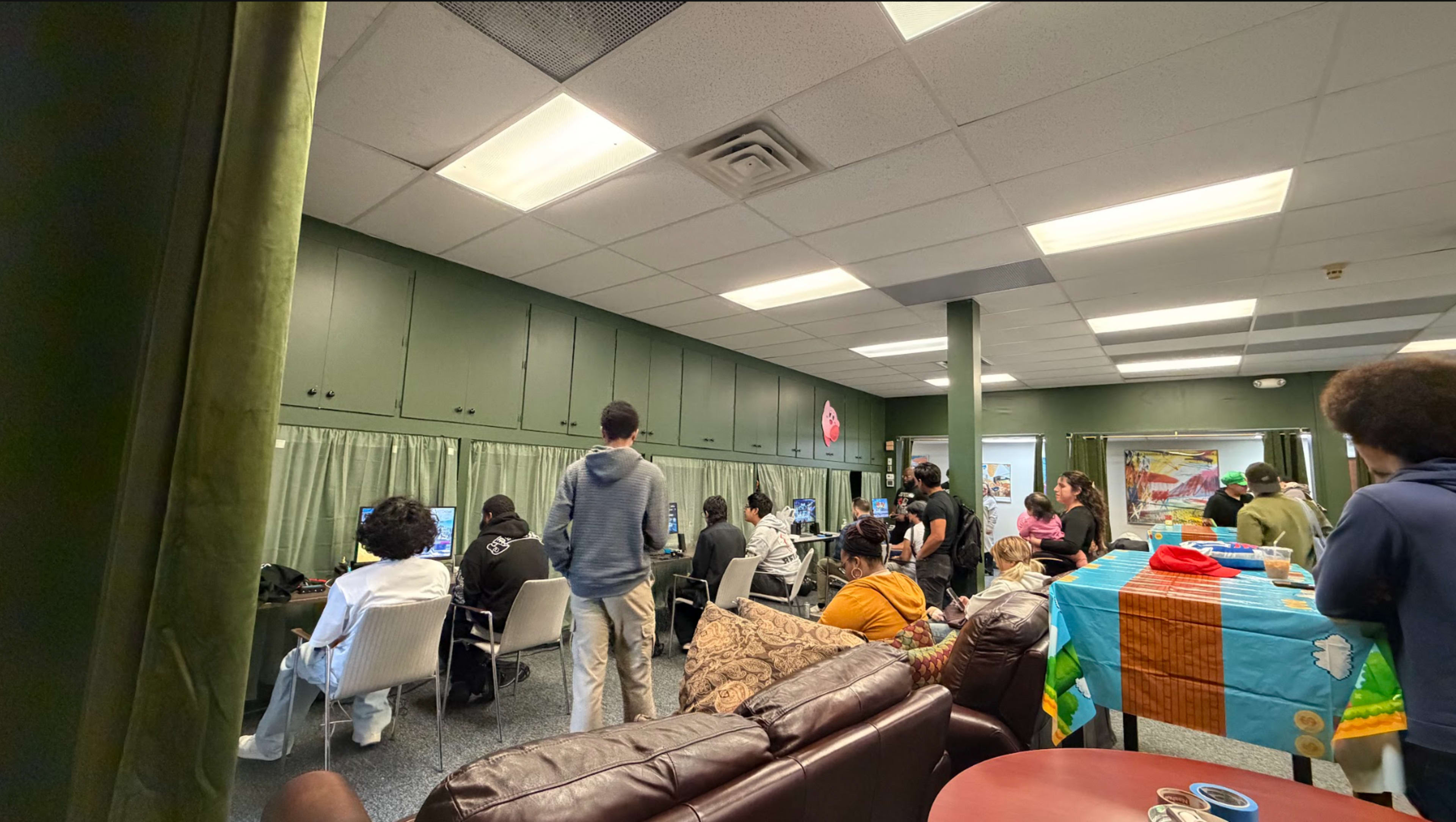 The image shows a room filled with people engaged around computer stations and a table with colorful tablecloths, while others gather nearby.