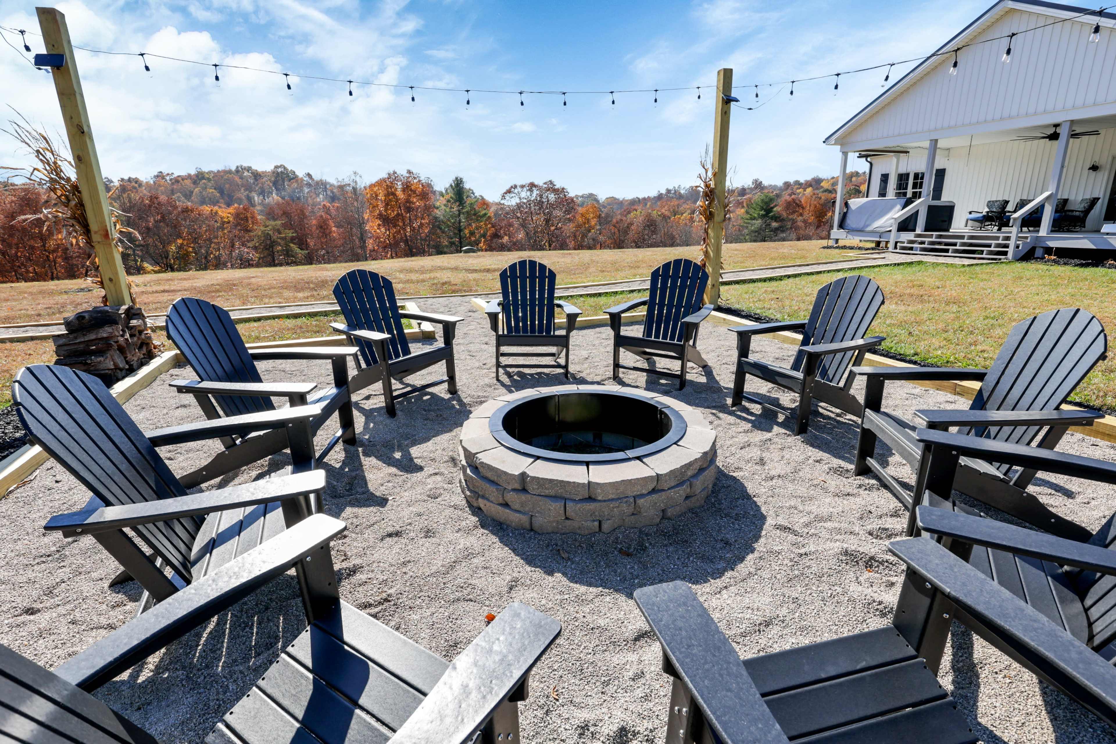 A circular seating area with black Adirondack chairs surrounds a stone fire pit, set on gravel with a view of autumn foliage in the background.