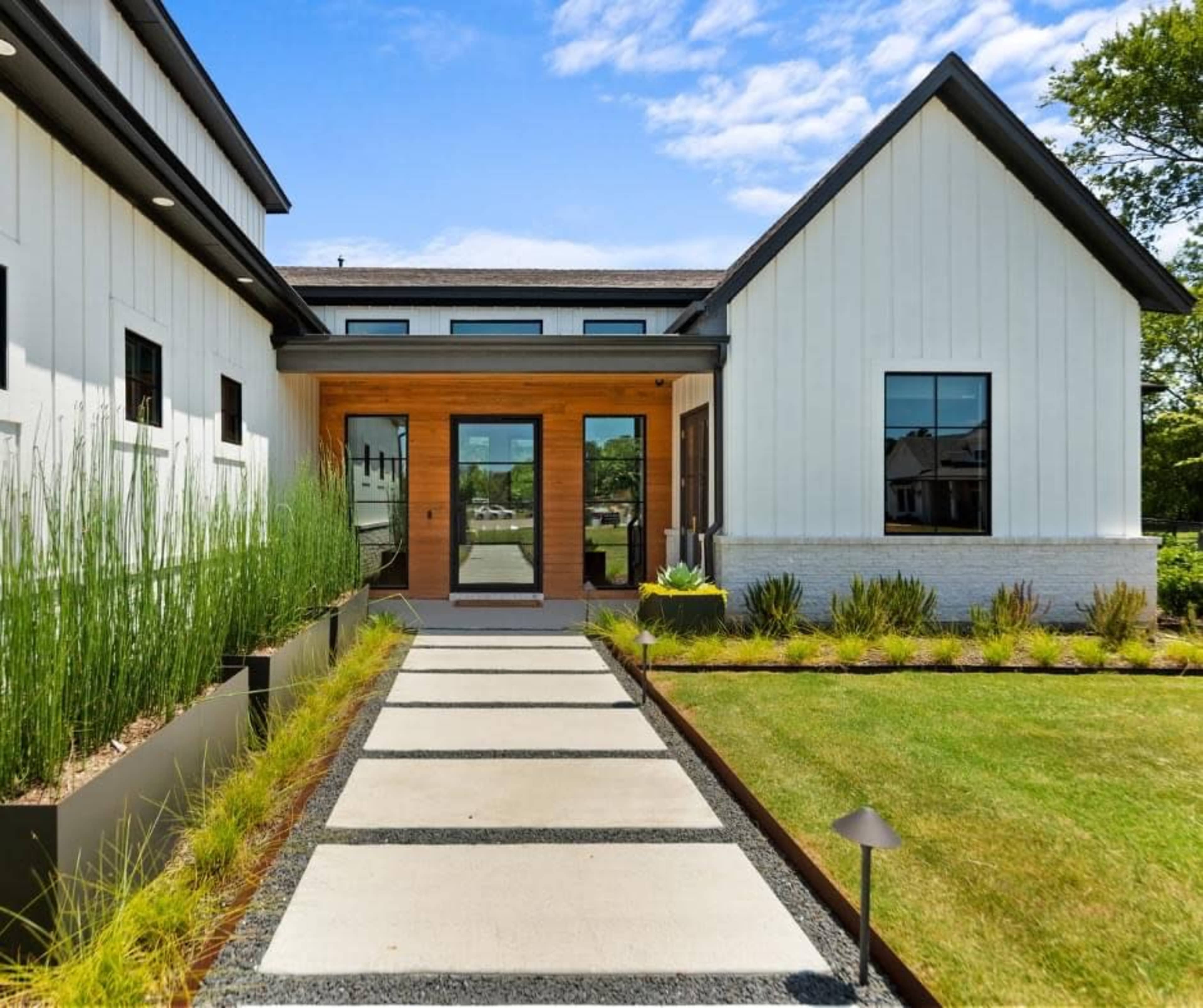 The image shows a modern home entryway with a stone walkway leading to large wooden double doors, bordered by neatly arranged grasses and shrubs.