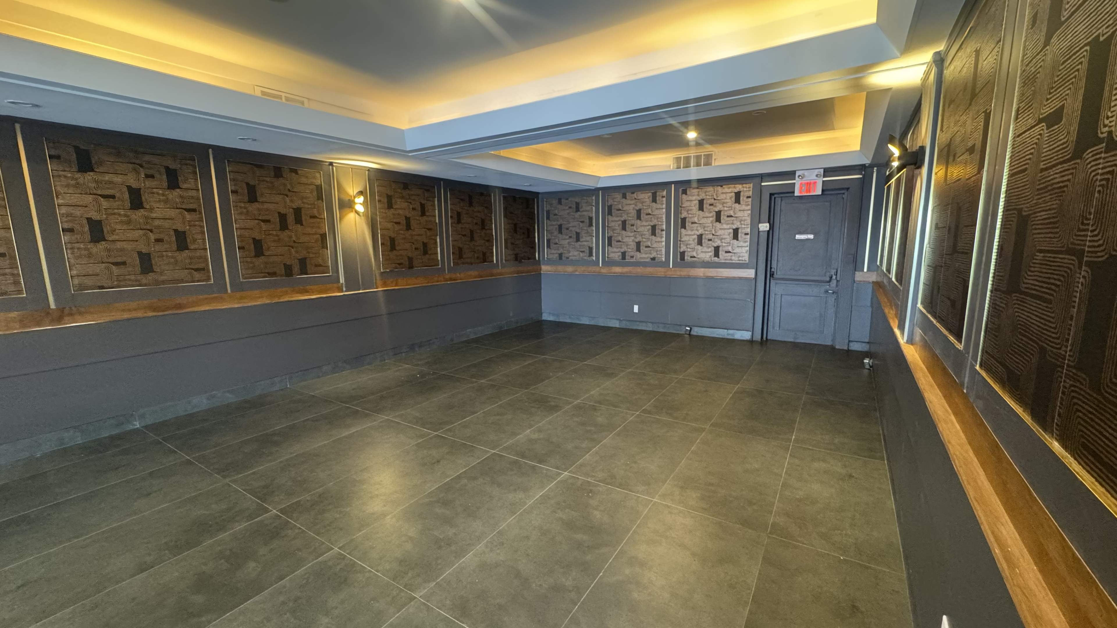 The image shows an empty room with dark tiled flooring, textured wall panels, and a single door.