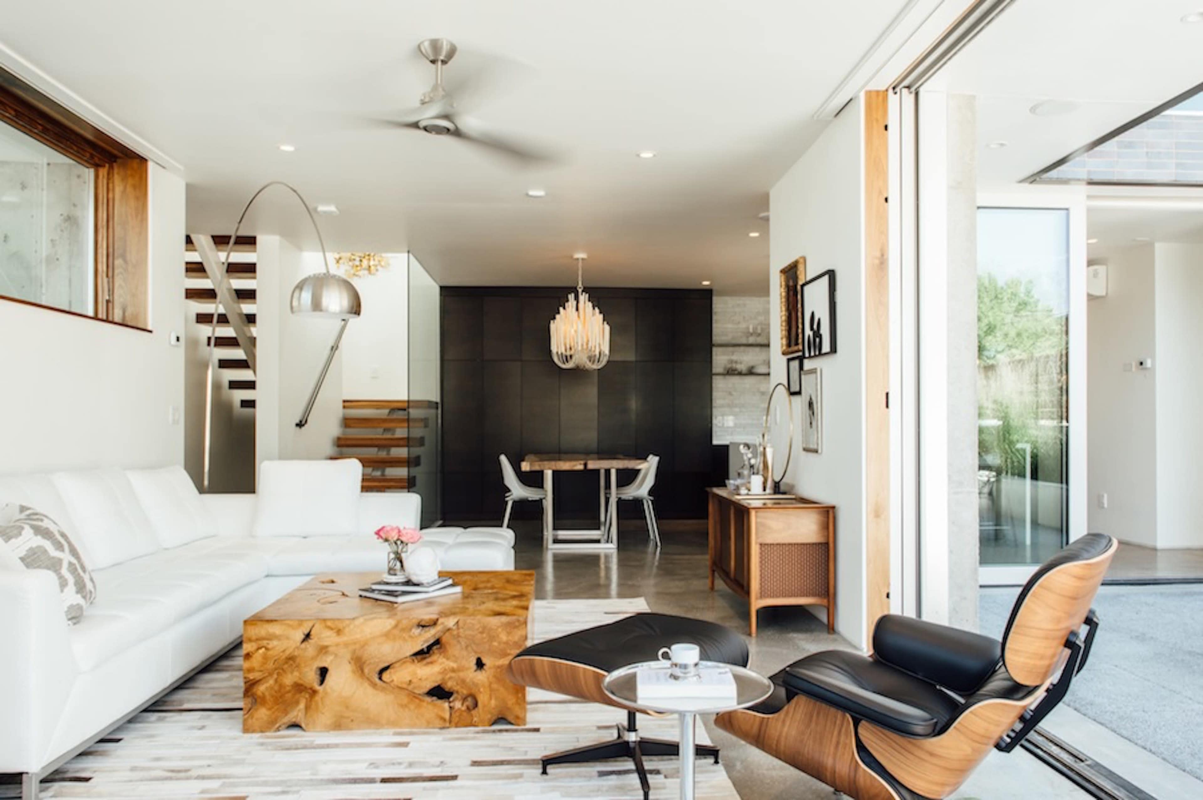 The image shows a modern living room featuring a white sectional sofa, a wooden coffee table, a sleek lounge chair, and an open layout connected to a dining area.