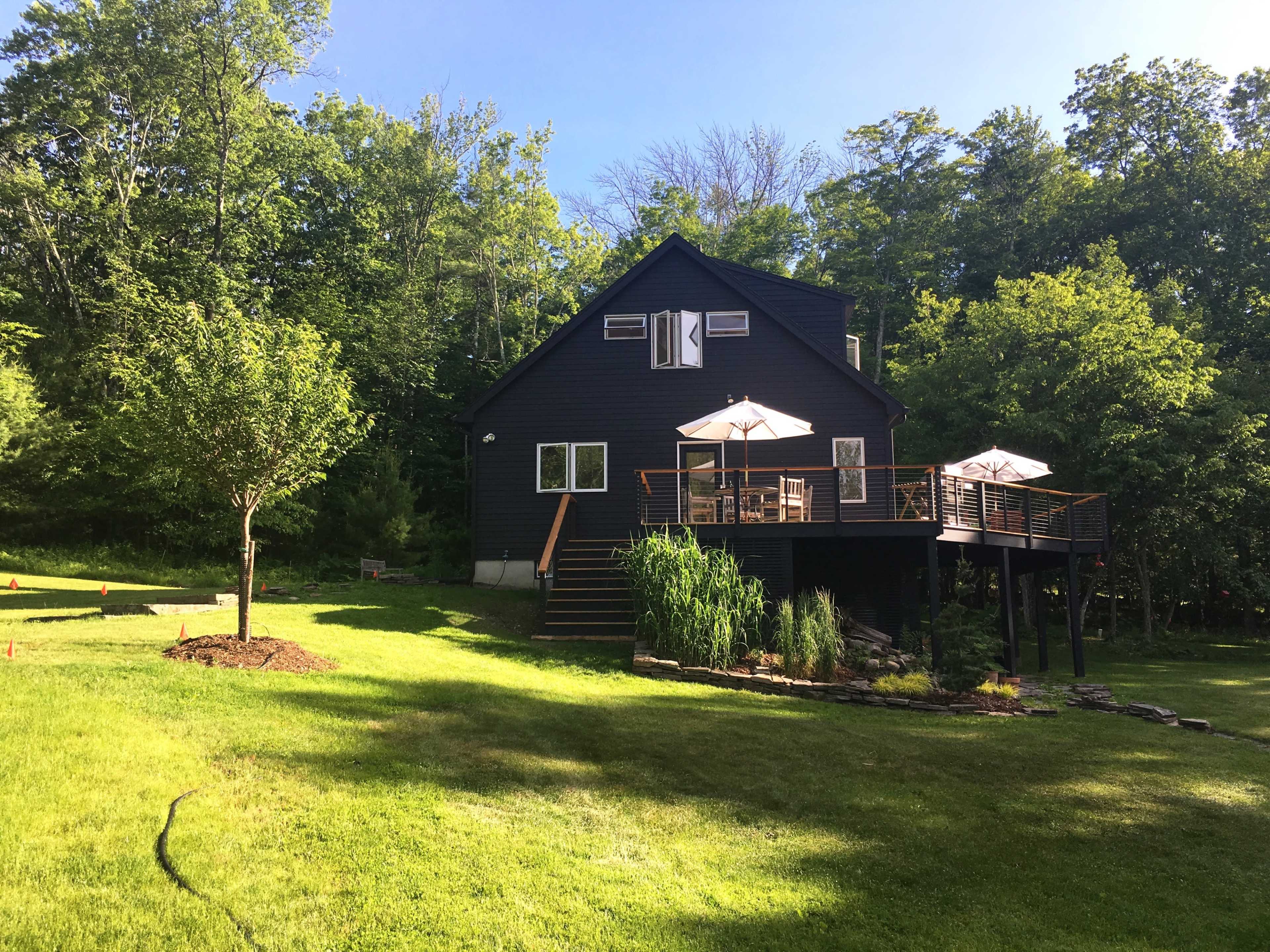 A black wooden house with a deck and patio umbrellas stands in a lush green yard surrounded by trees.