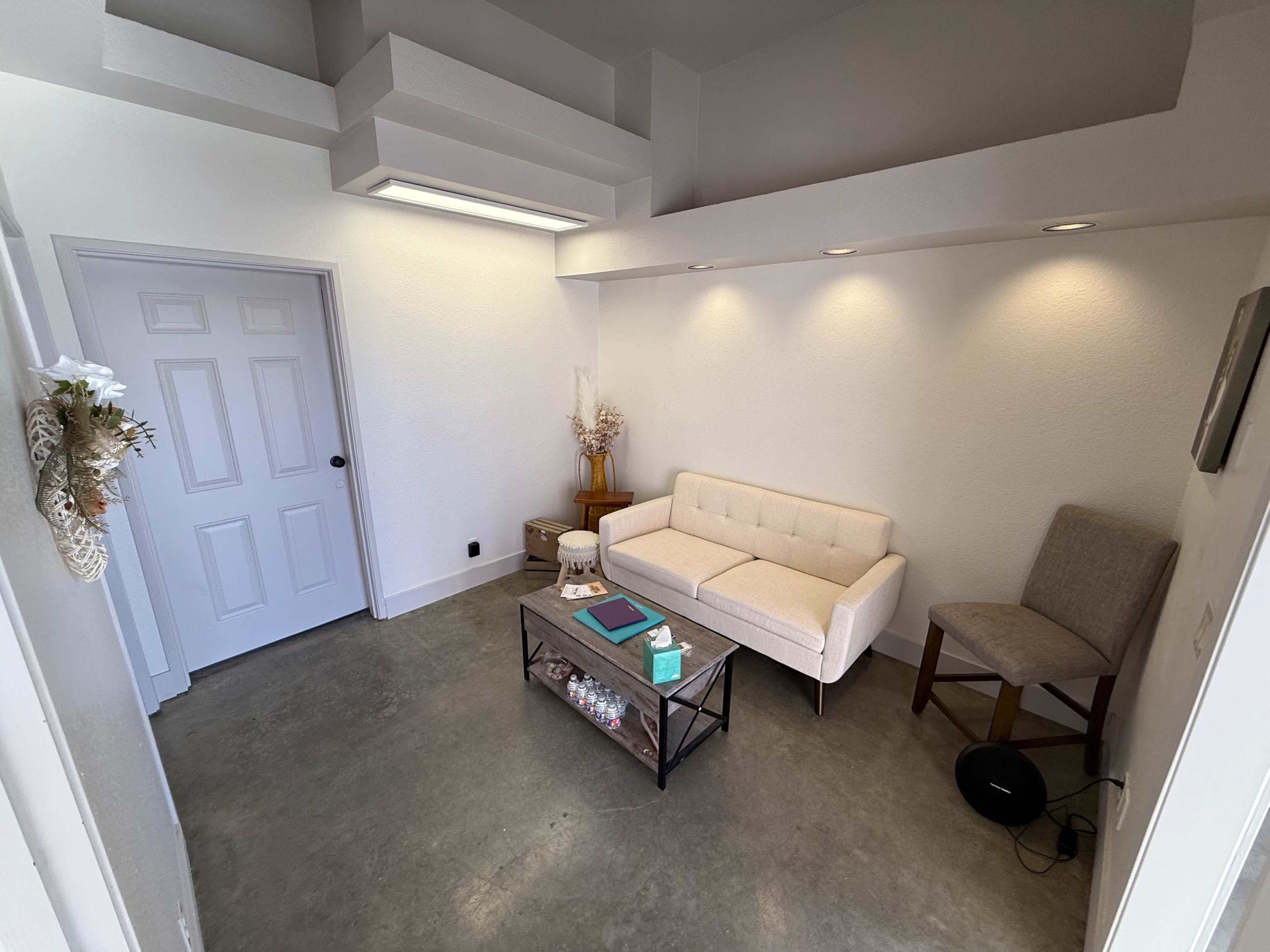 The image shows a modern waiting area featuring a white couch, a glass coffee table with decorative items, and a single chair in a well-lit space with neutral-colored walls.