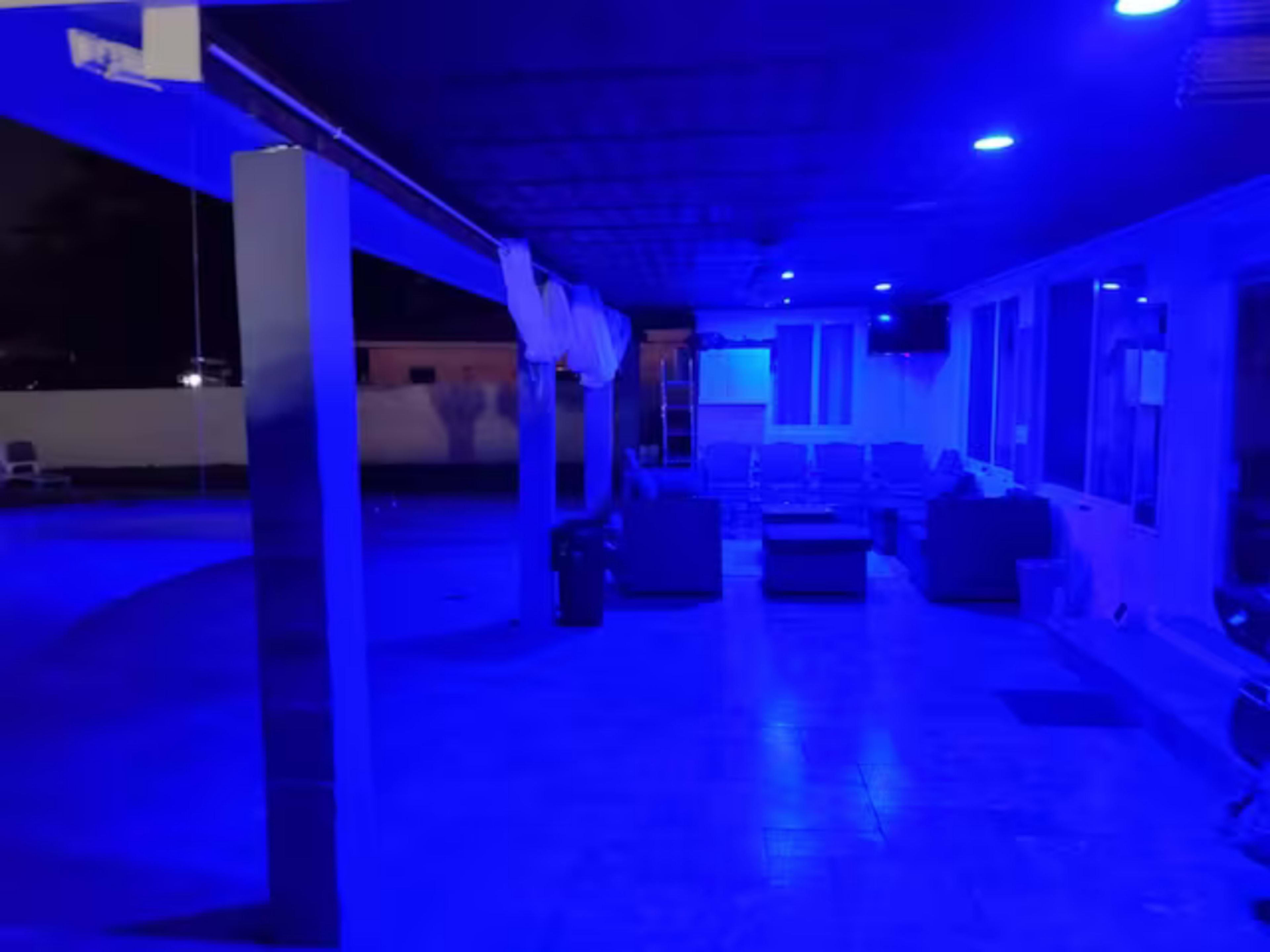 The image shows a poolside area illuminated by blue lighting, featuring lounge furniture and a covered seating space.