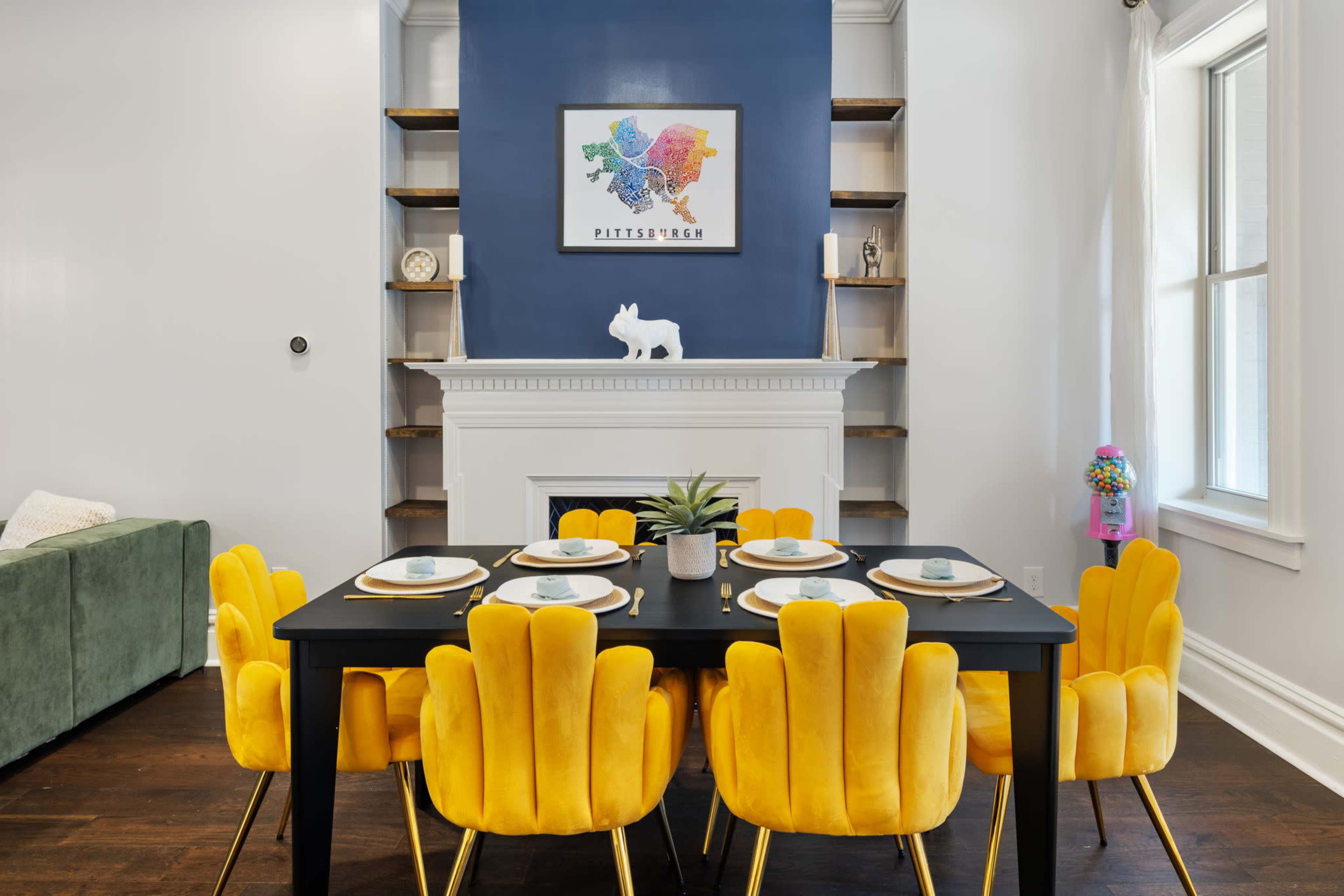 A dining area features a black table set with yellow chairs, against a blue wall displaying a framed map artwork above a white fireplace.