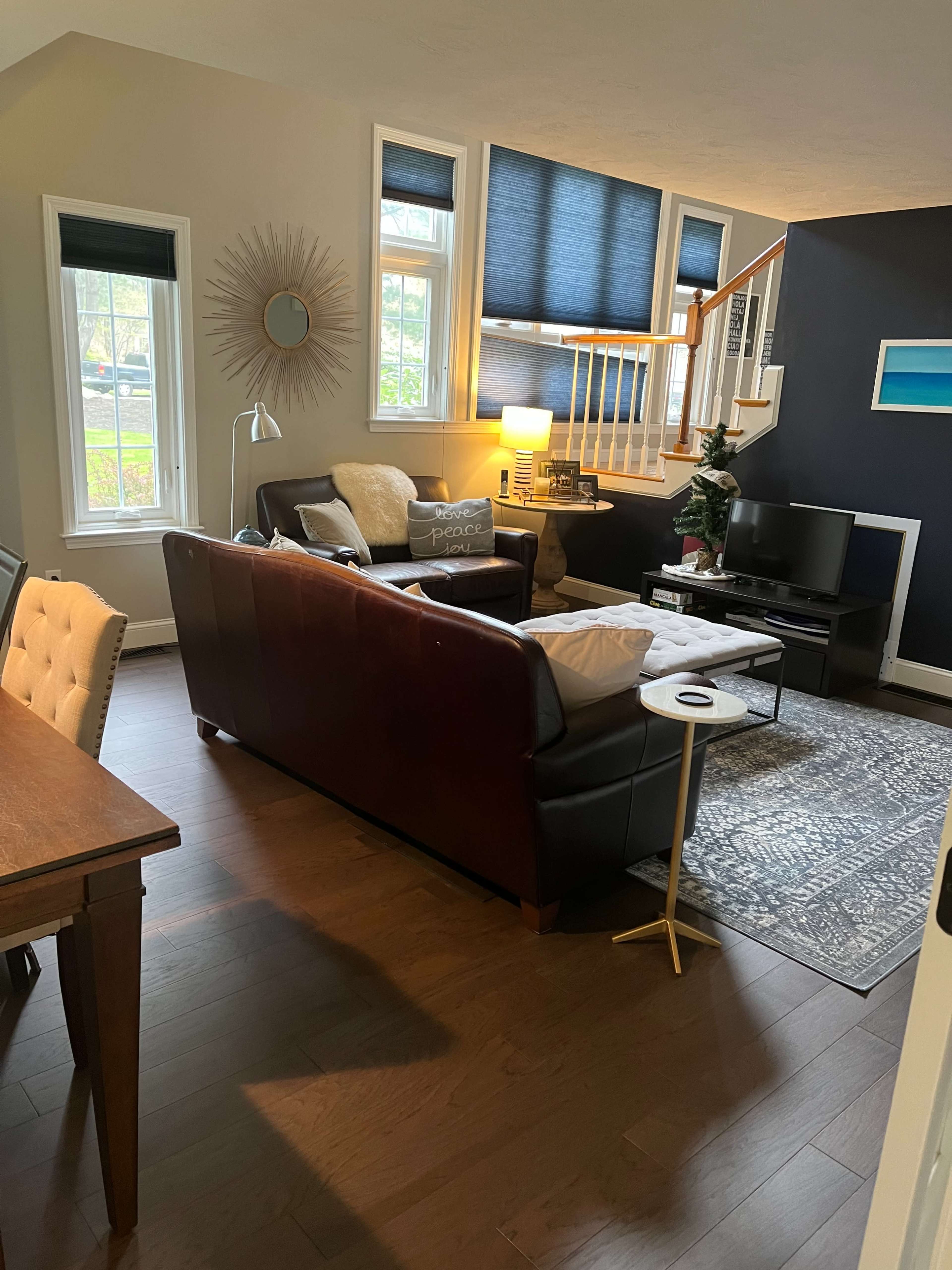 The image shows a cozy living room with a dark sofa, a light-colored armchair, a coffee table, and large windows letting in natural light.