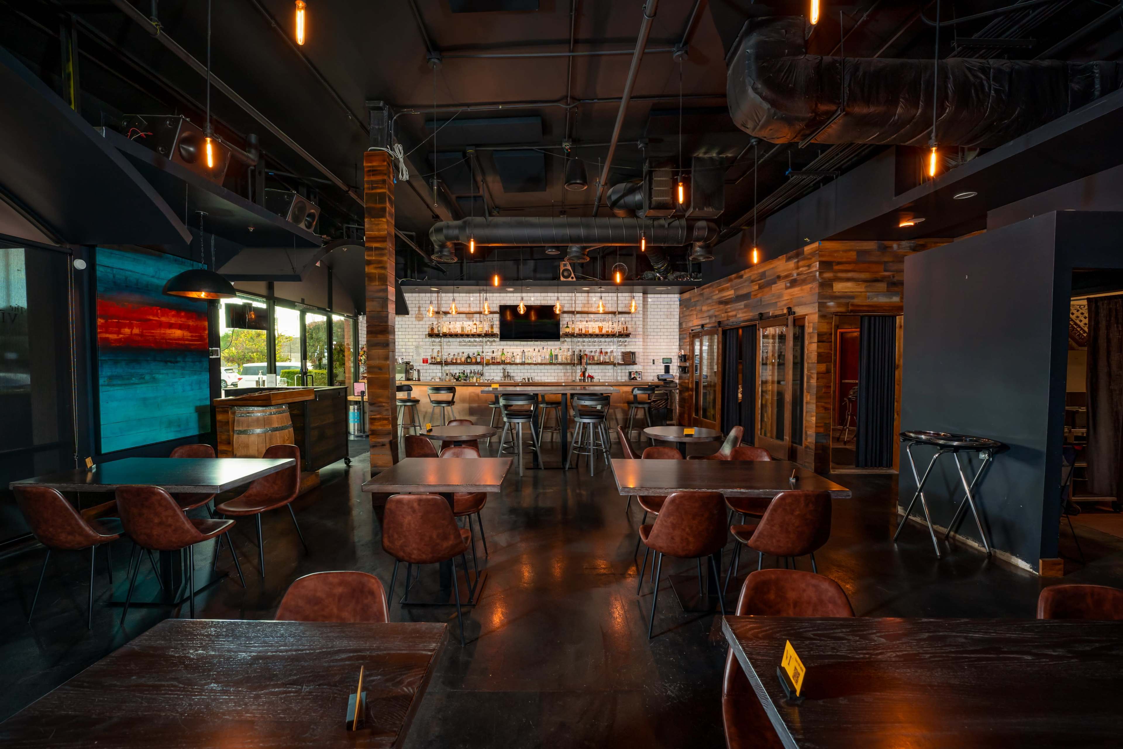 The image shows a modern restaurant interior featuring wooden tables, leather chairs, and a bar with shelves of liquor in the background.