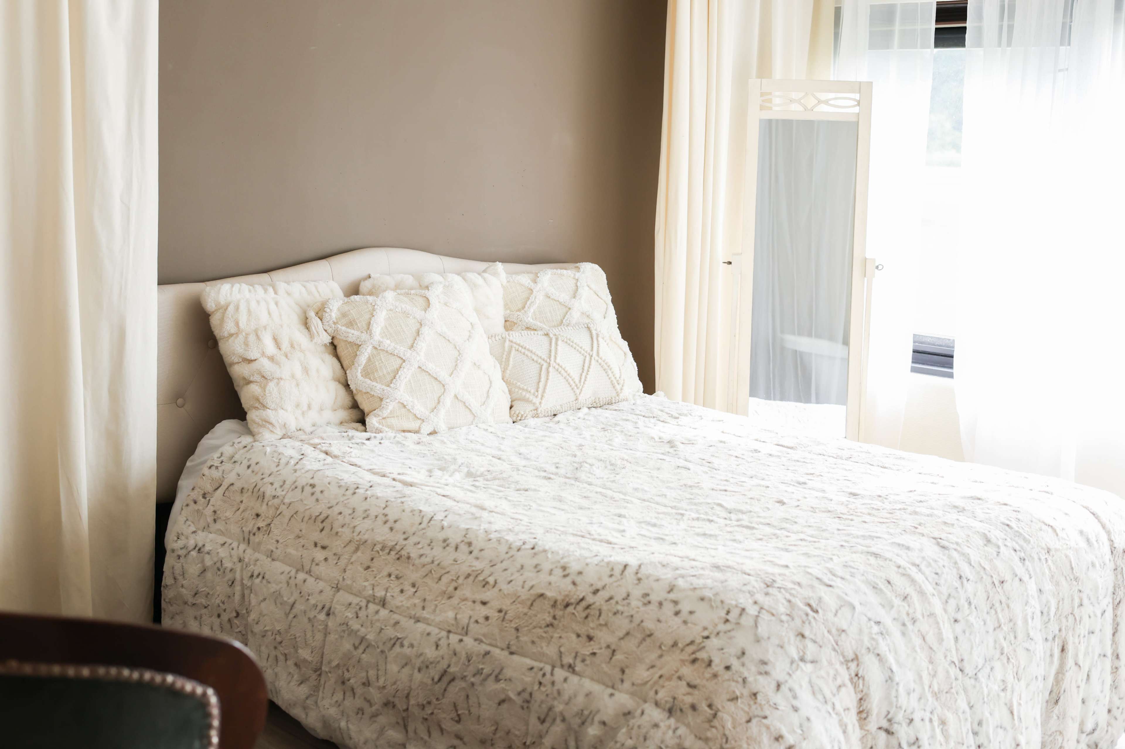 A neatly made bed with a light-colored blanket and decorative pillows is positioned against a wall in a bright bedroom, accompanied by a full-length mirror and sheer curtains.
