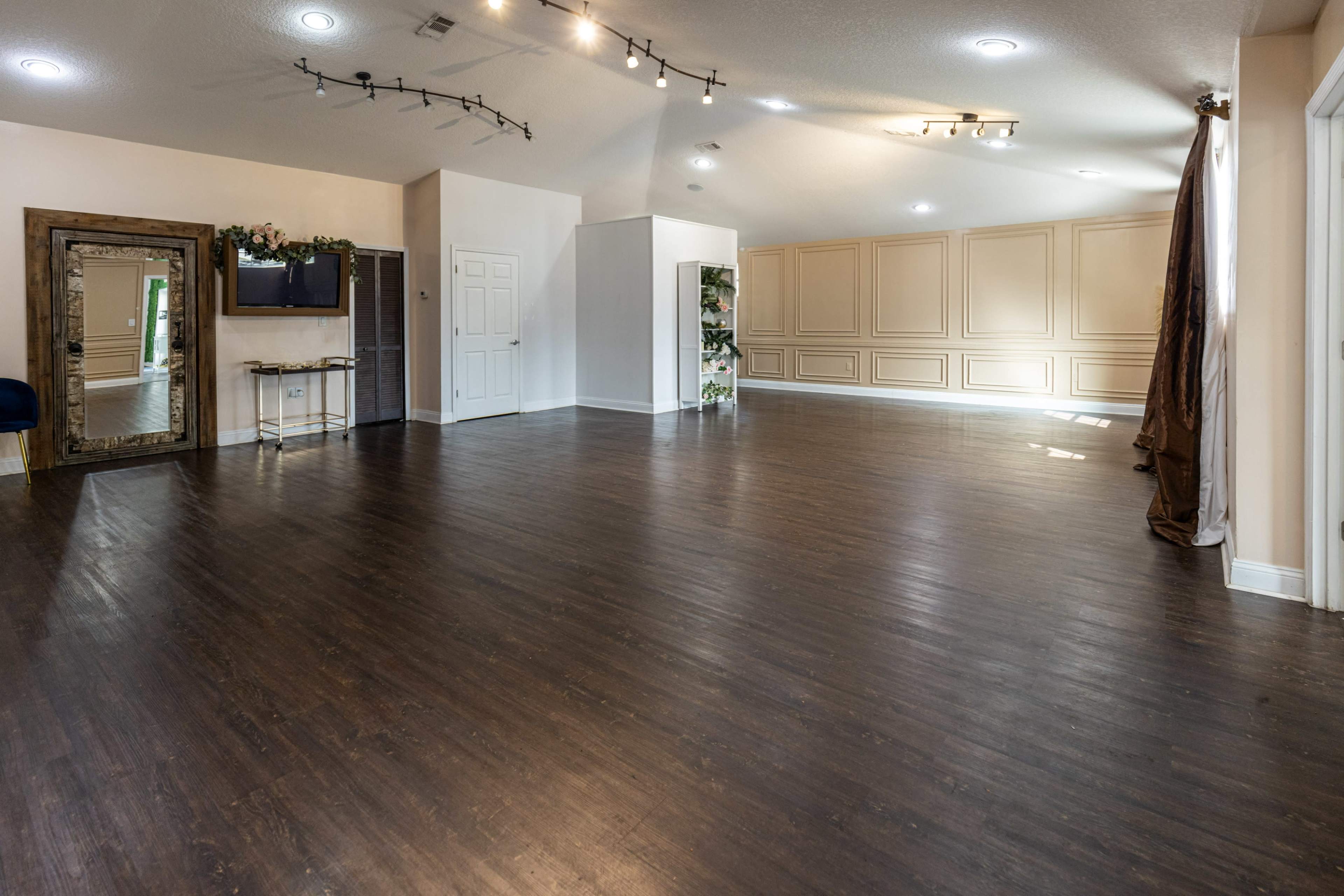 A spacious, empty room with dark wooden flooring, a large mirror, and light-colored walls featuring decorative panels.
