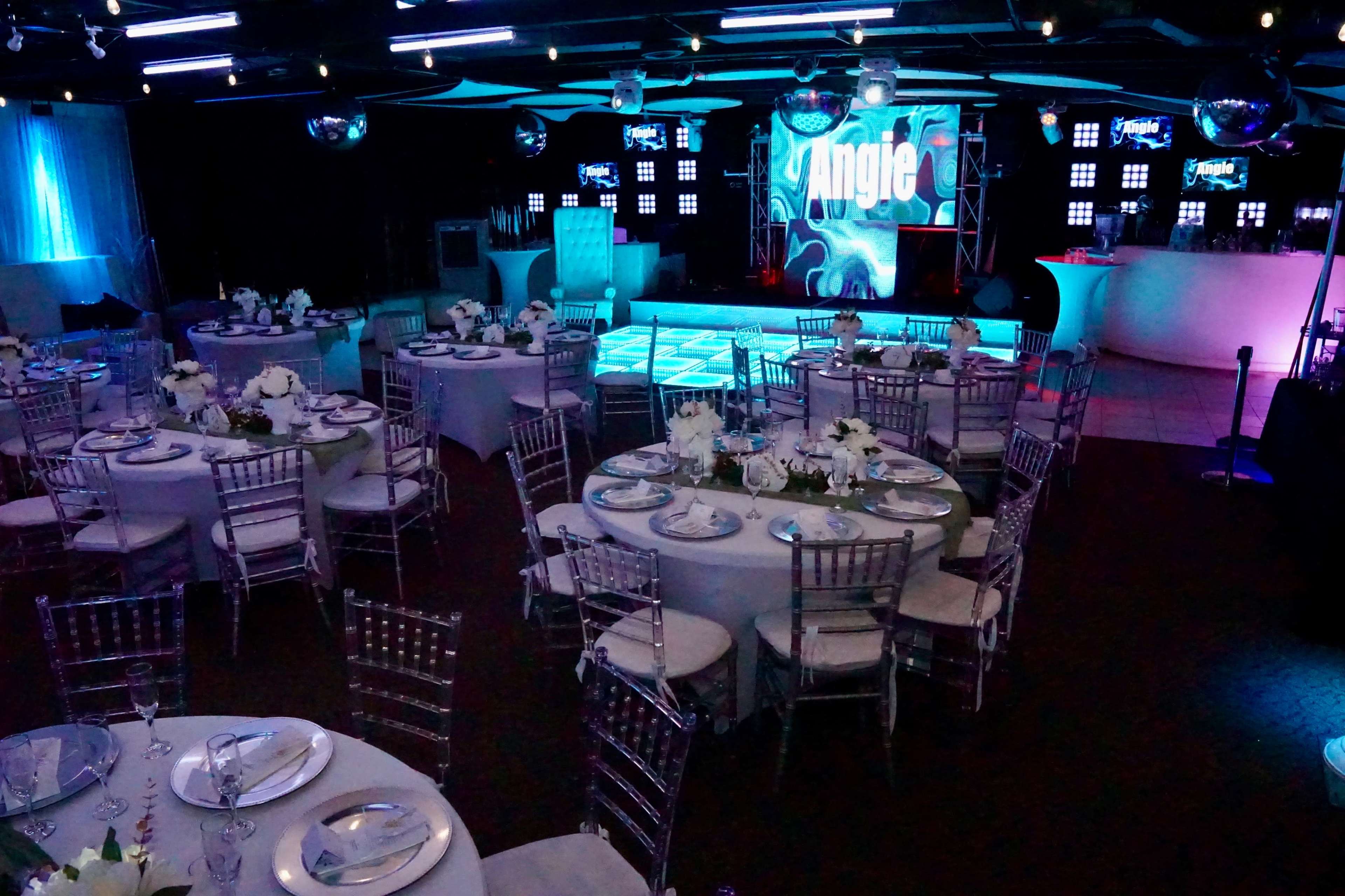 A banquet hall set up for an event, featuring circular tables with white tablecloths and floral centerpieces, a lit dance floor, and a large screen displaying the name "Angie."
