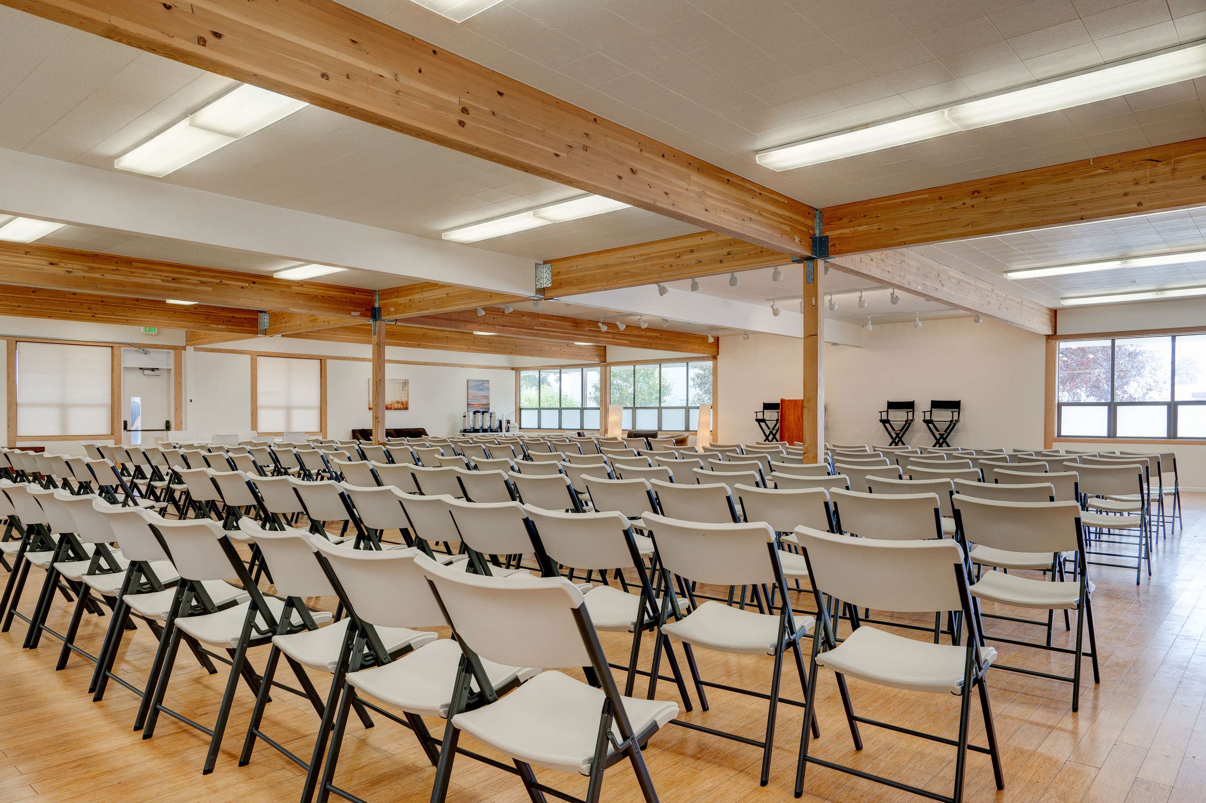 A spacious indoor venue features rows of empty folding chairs arranged for an event, with wooden beams and large windows providing natural light.