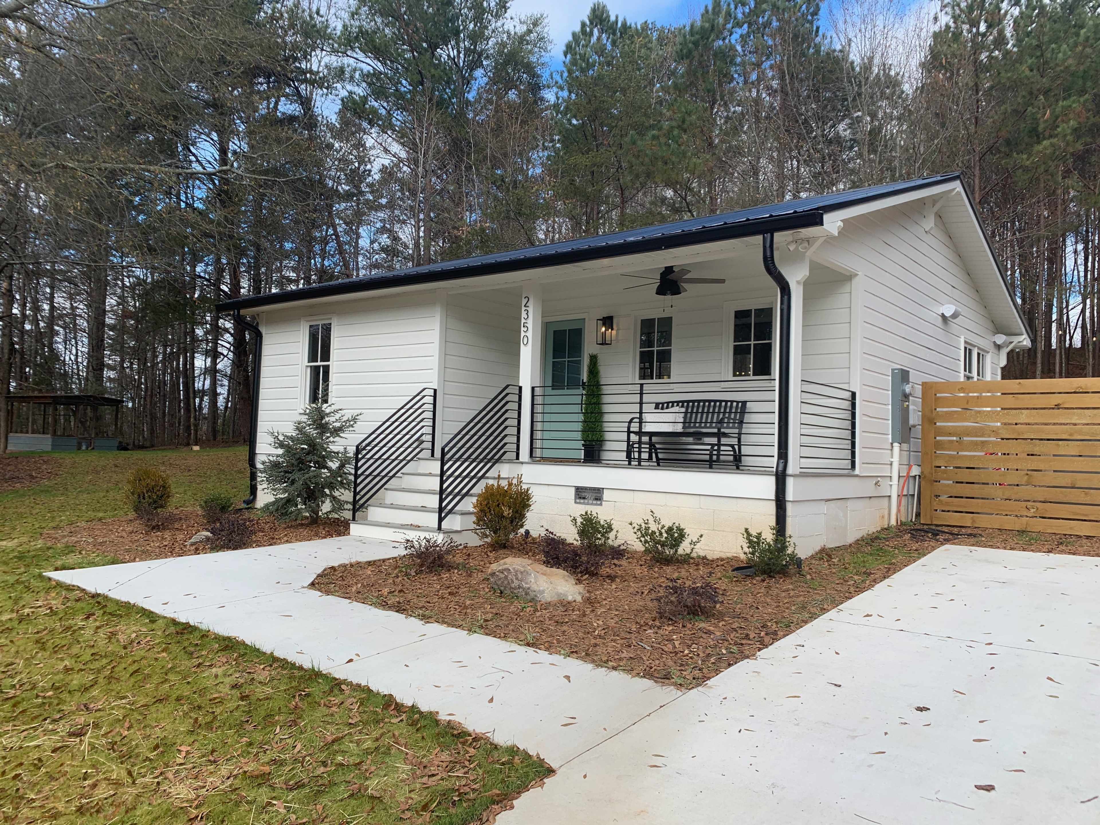 A modern single-story white house with a black roof, featuring a front porch and surrounded by landscaped greenery.