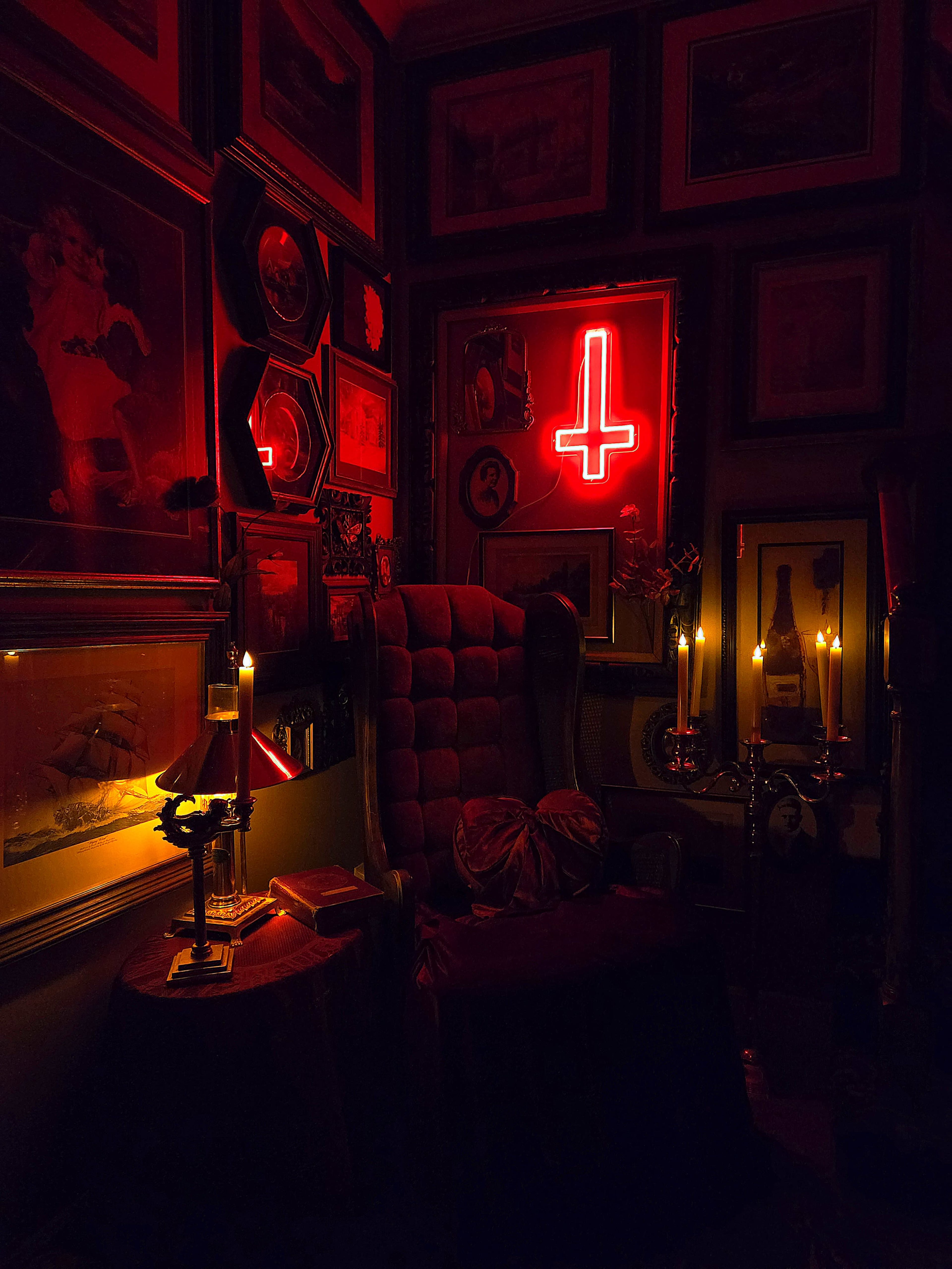 The dimly lit room features a large, ornate armchair surrounded by eclectic framed artwork and illuminated by candles and a red neon cross.