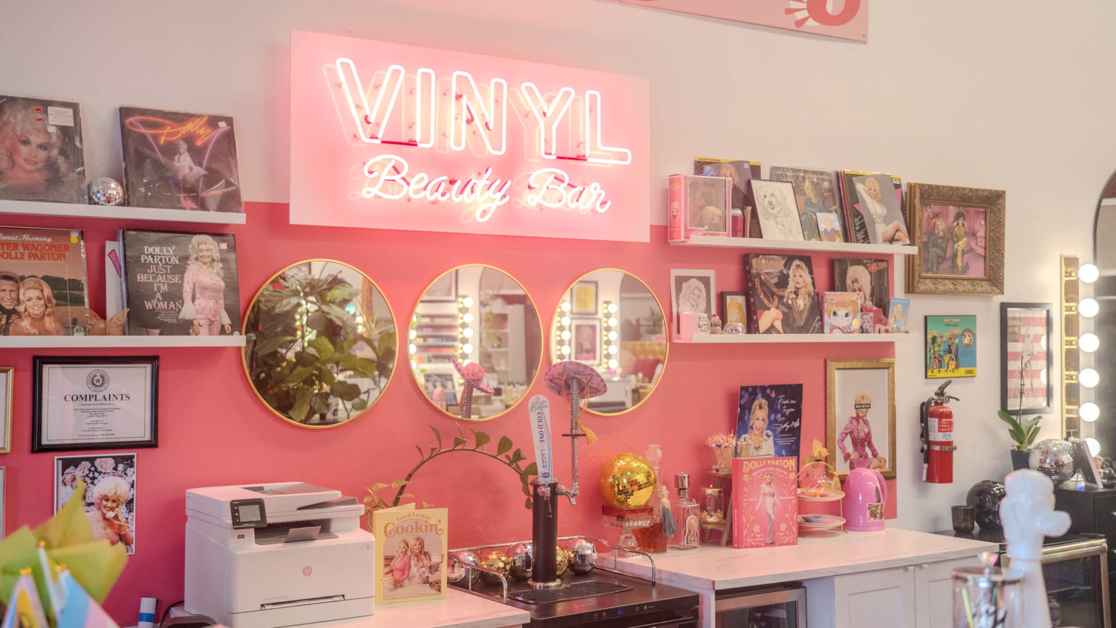 The image shows a brightly lit beauty bar with a neon "VINYL" sign, mirrors, shelves displaying various beauty products and decor, and a pink-themed interior.