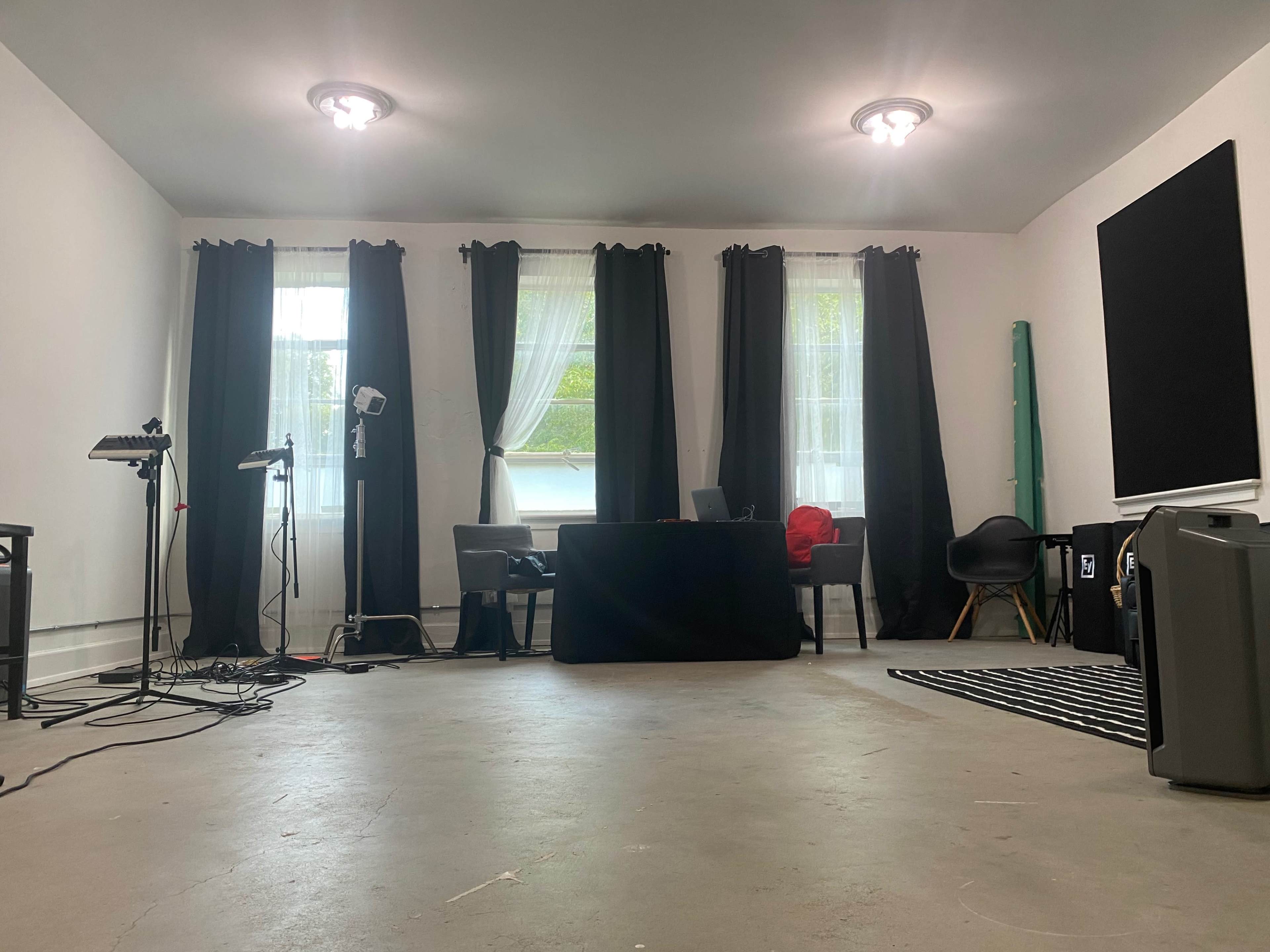 A spacious room with a table and chairs positioned near a window, surrounded by black curtains, and various studio equipment set up along the walls.