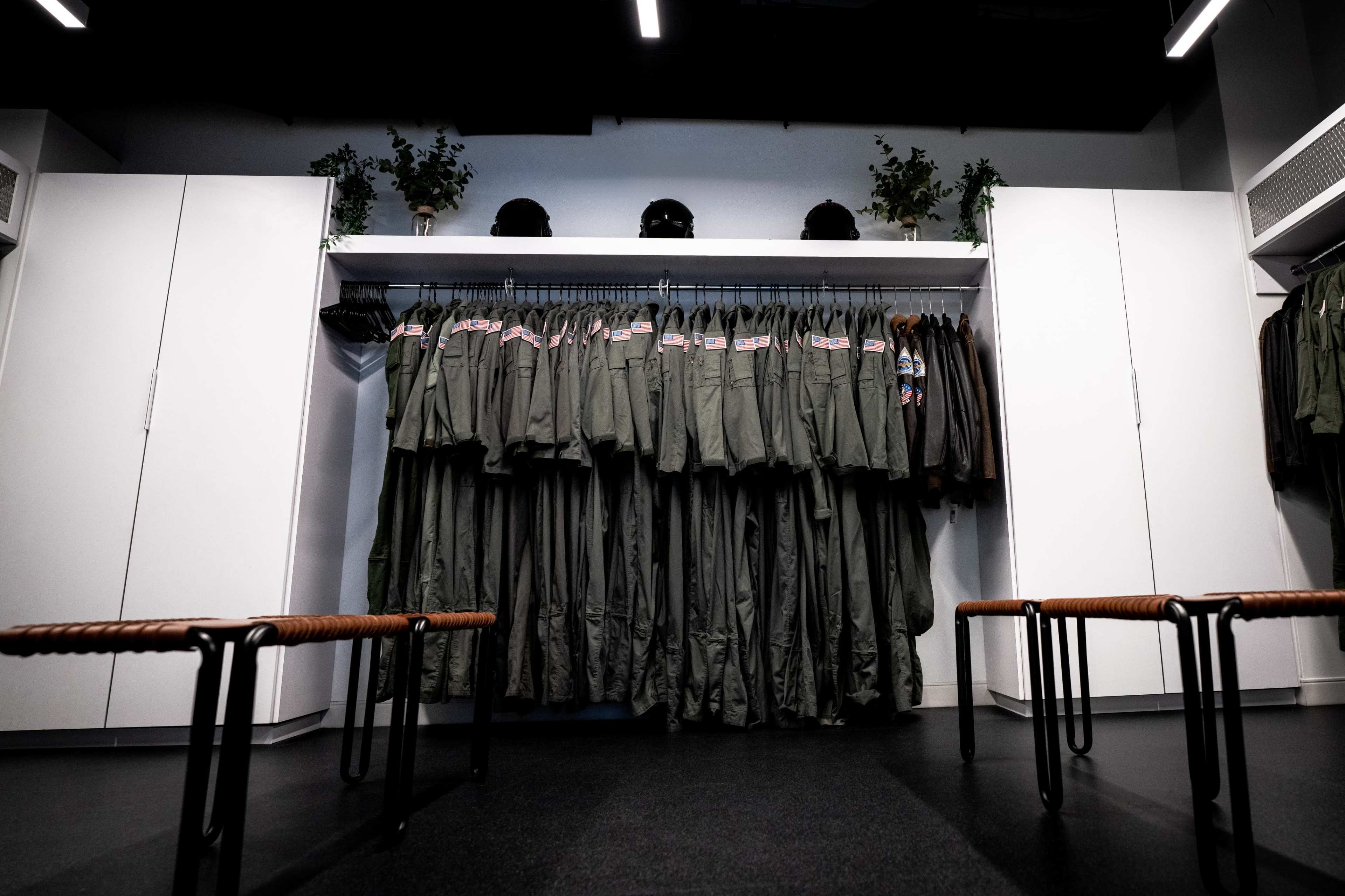 The image shows a row of military-style jumpsuits hanging on a rack in a neatly organized room with benches in the foreground.