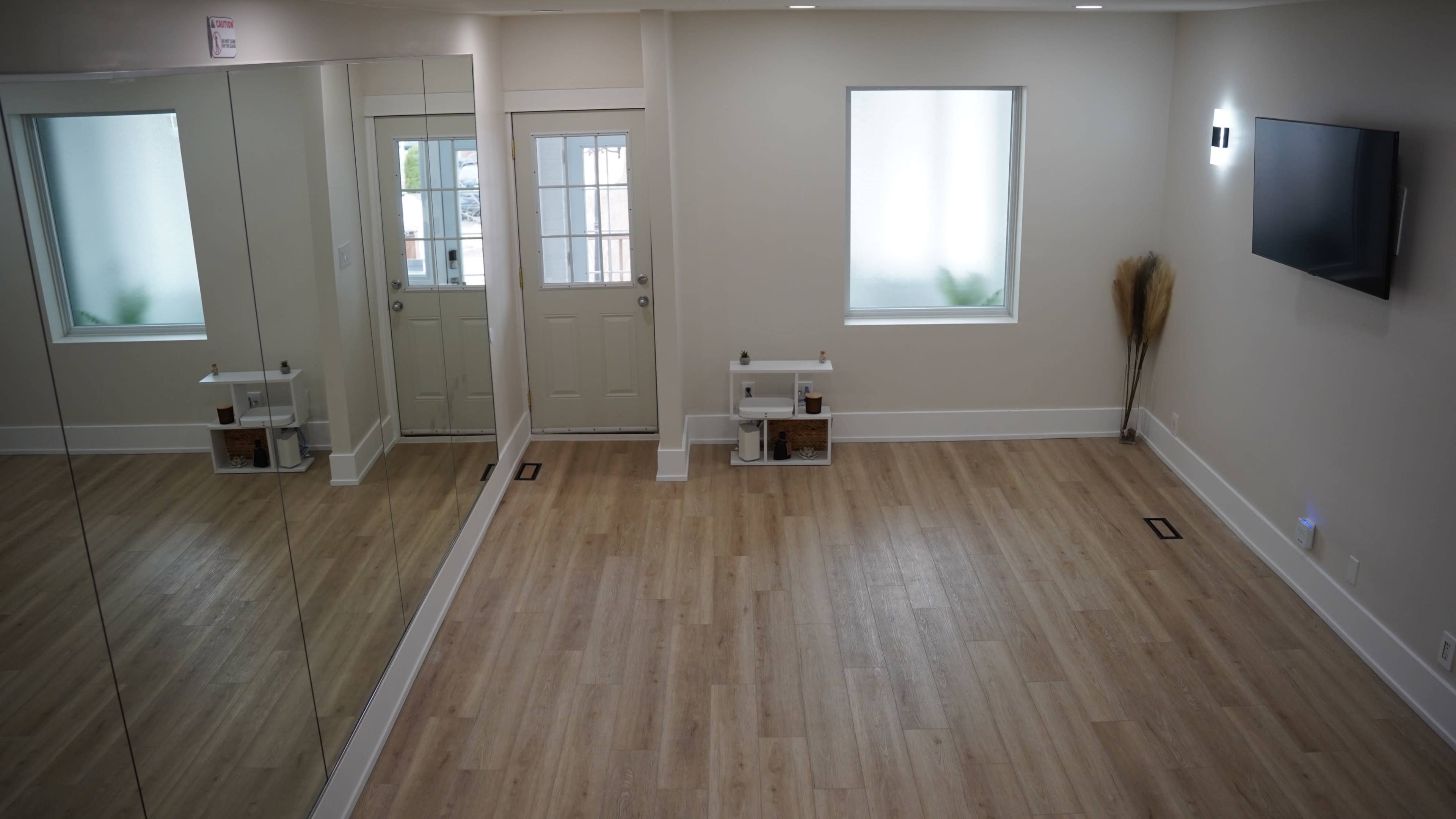 The image depicts a spacious, well-lit room with light wooden flooring, a large mirror on one wall, a door, and minimal furnishings, including a small table and a decorative plant.