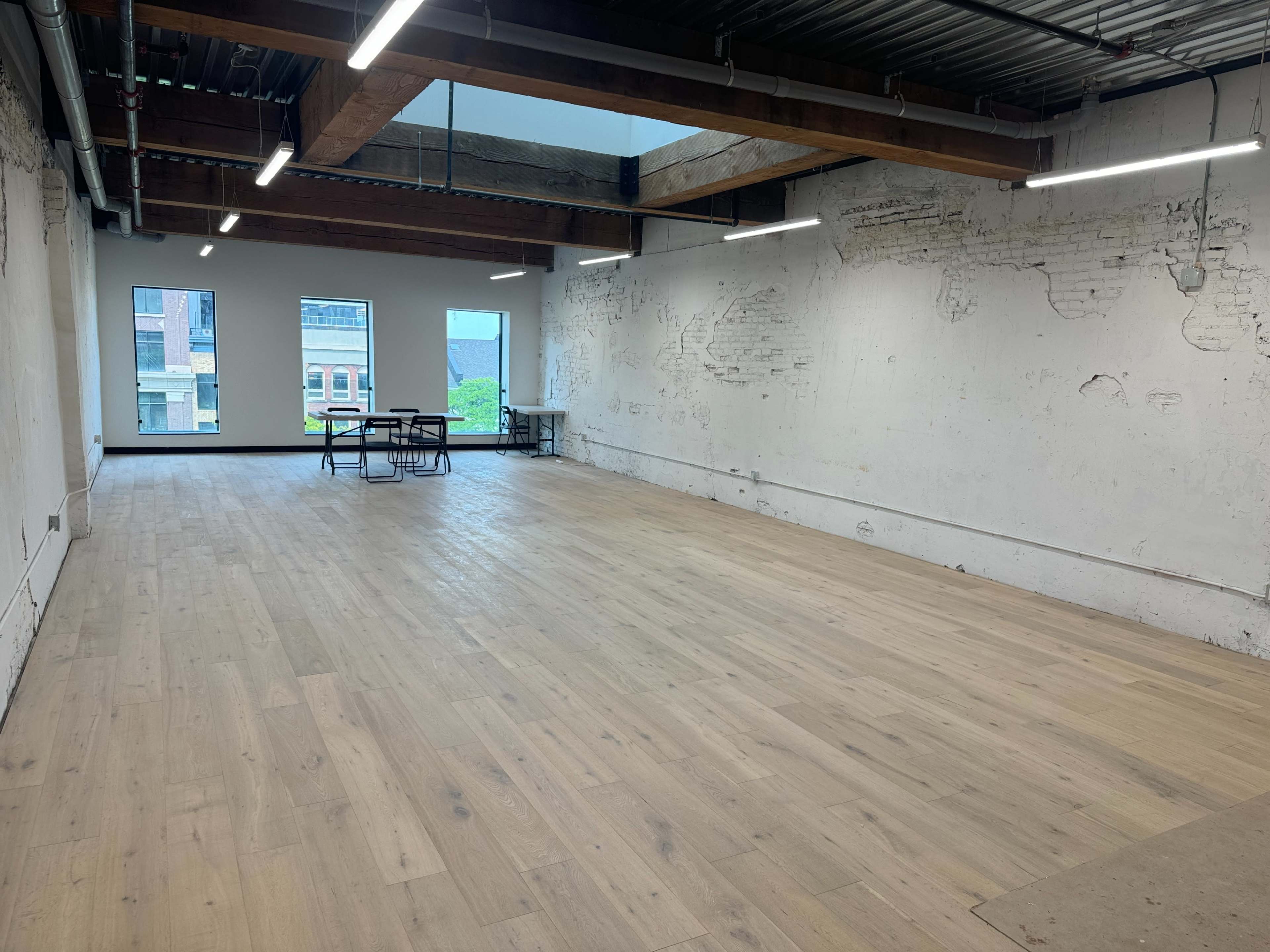 A spacious, unfinished interior room features large windows and wooden beams overhead, with a few tables positioned in the center and bare walls.