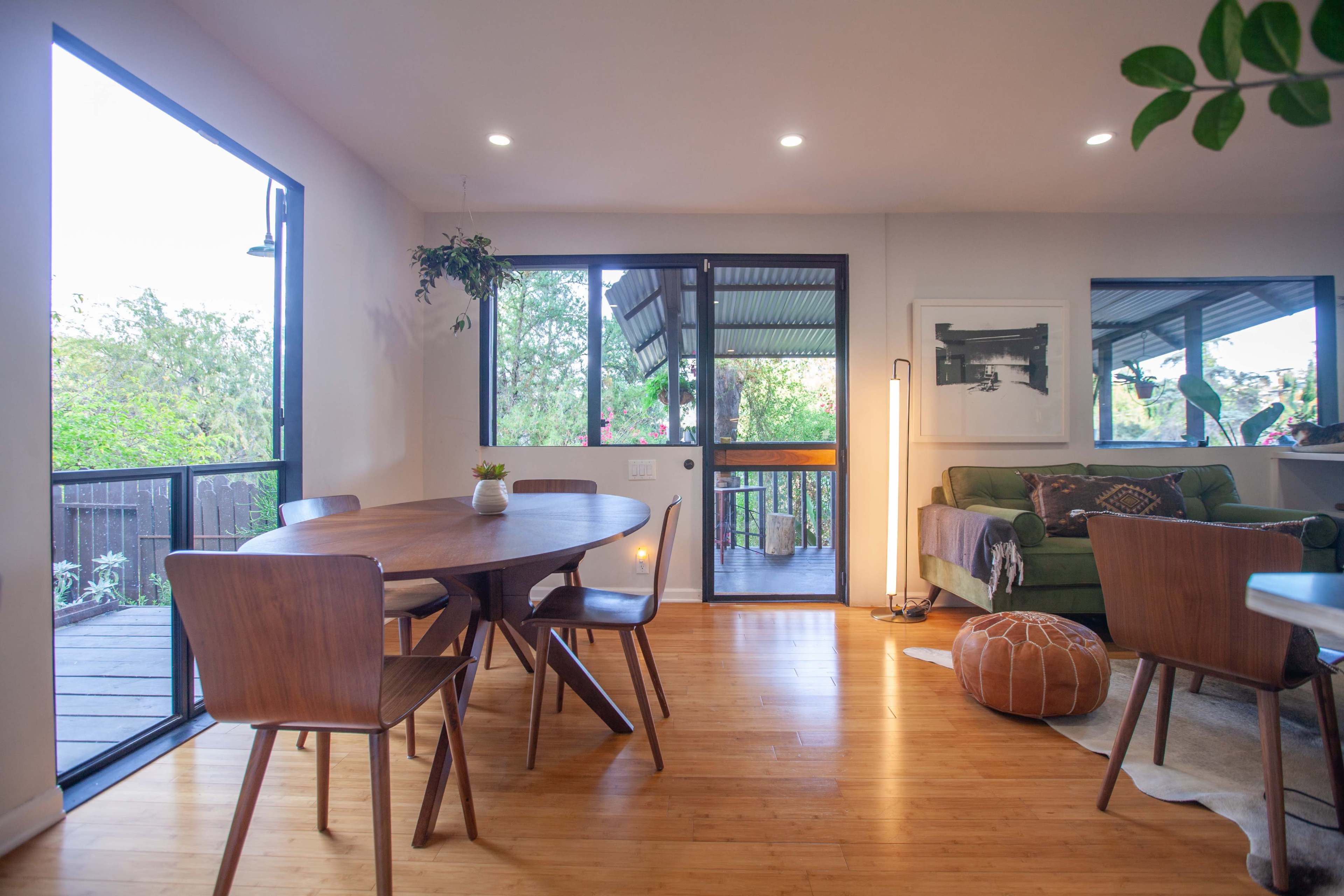 A modern living space with a wooden dining table and chairs, a green sofa, and large windows allowing natural light in.