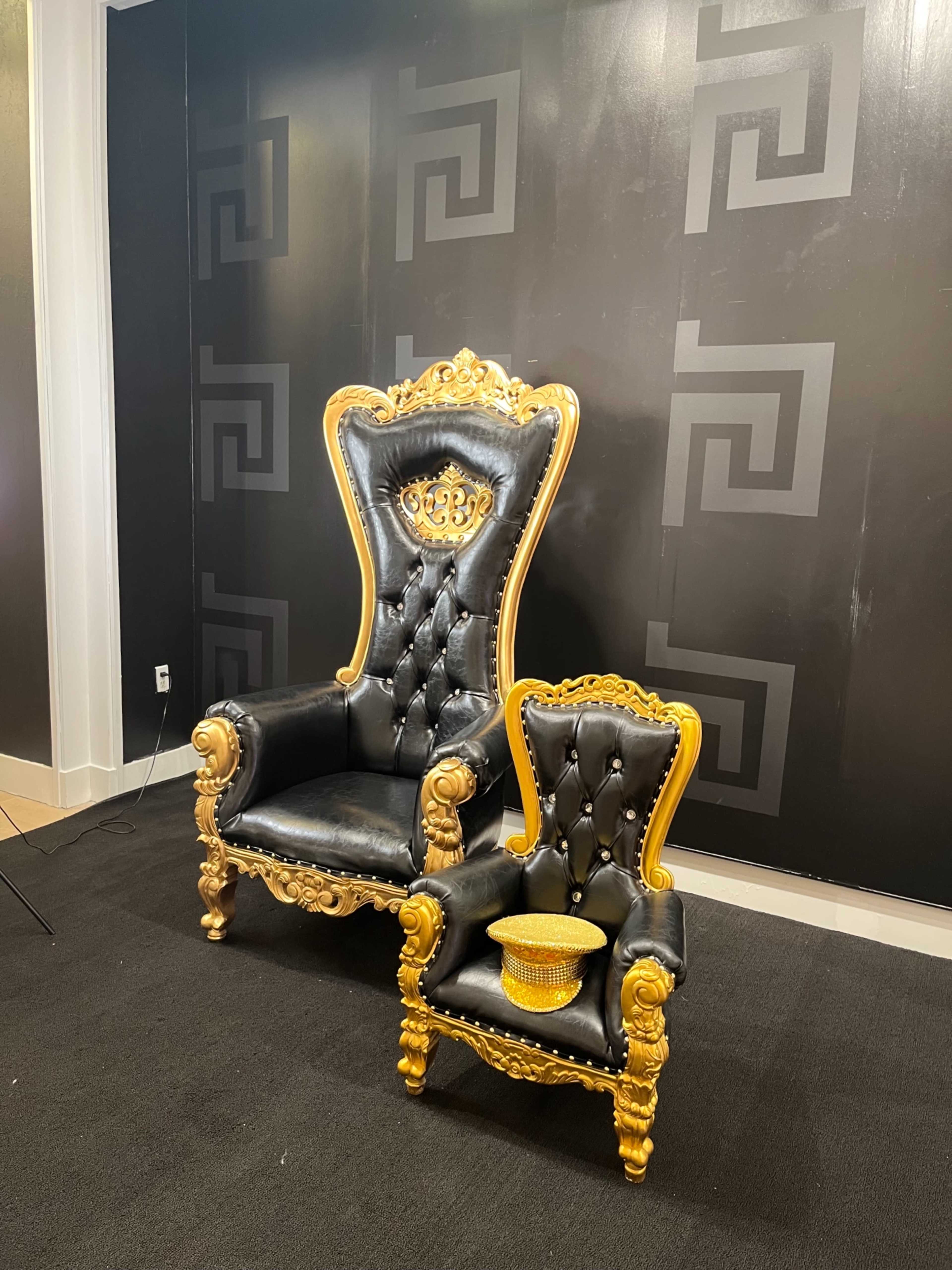 A pair of ornate black and gold Victorian-style chairs, with one large chair and a smaller matching chair beside it, set against a dark wall featuring a repeating geometric pattern.