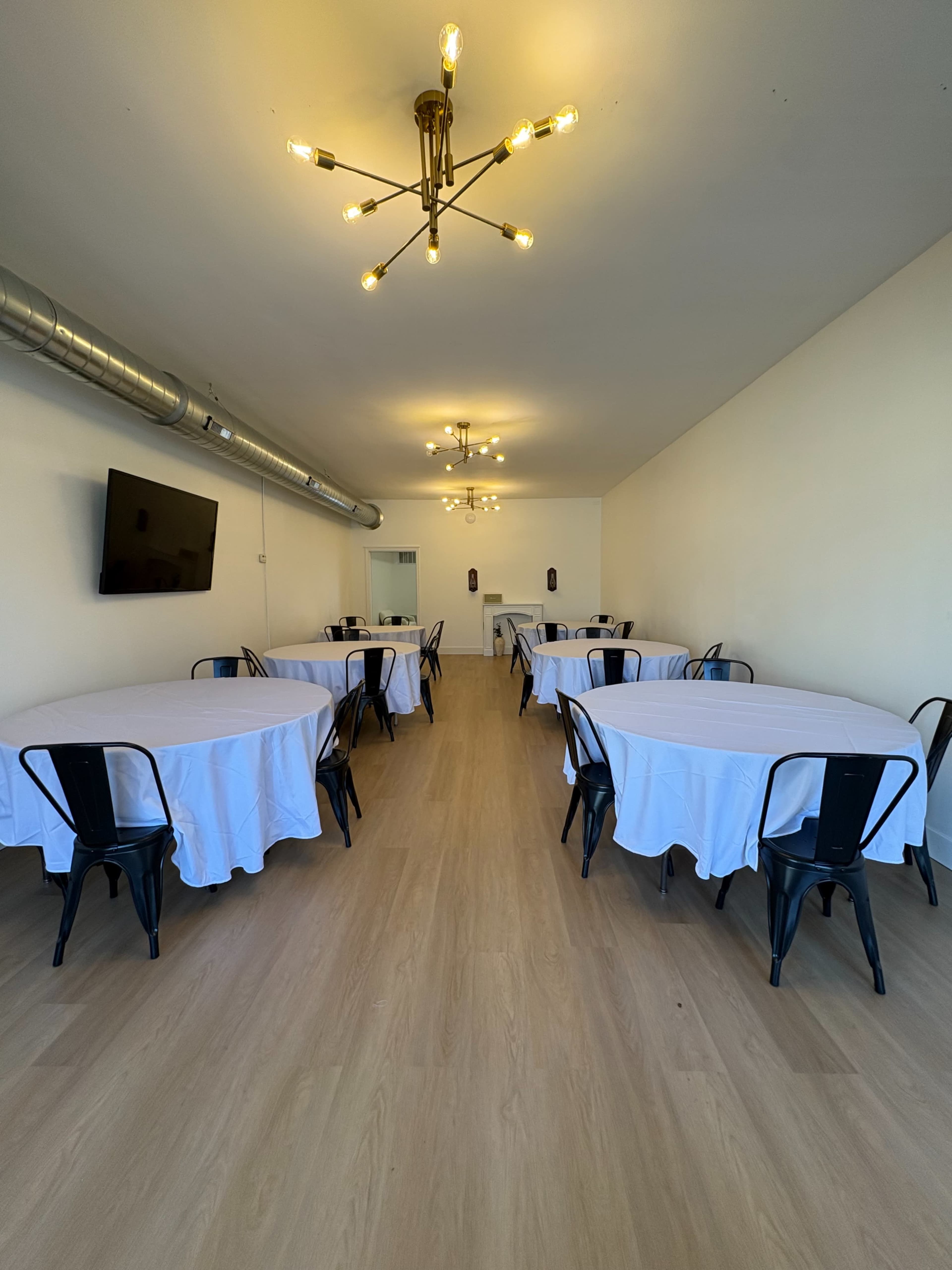 The image shows a spacious room with several round tables covered in white tablecloths, black chairs, and light fixtures on the ceiling.